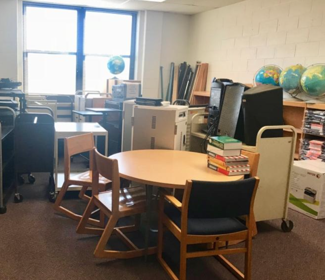 A small office or classroom with cluttered furniture including desks, chairs, globes, and stacks of books, illuminated by natural light from windows.