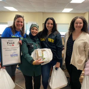 Four women standing together indoors, one holding a food donation, with a sign that reads 'Food Hub' and 'Food Drive'.