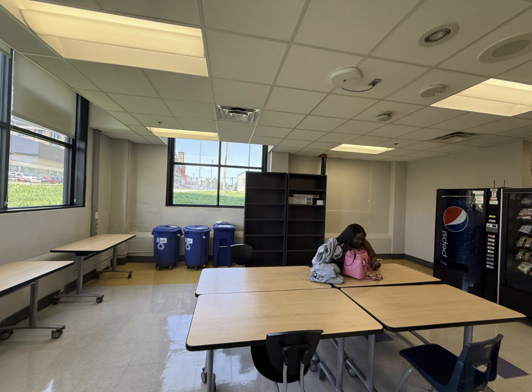 An empty break room or cafeteria with tables, chairs, a vending machine, recycling bins, and a woman sitting with a pink bag.