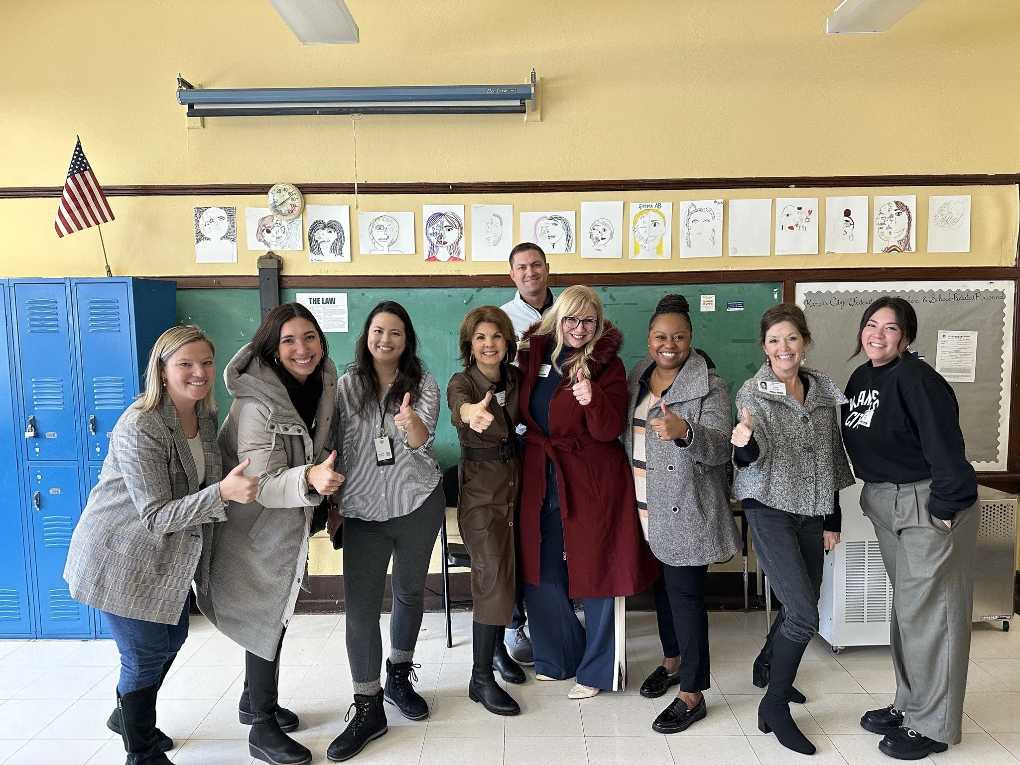 A diverse group of nine people standing in a school hallway, smiling and giving thumbs-up, with artwork and lockers in the background.