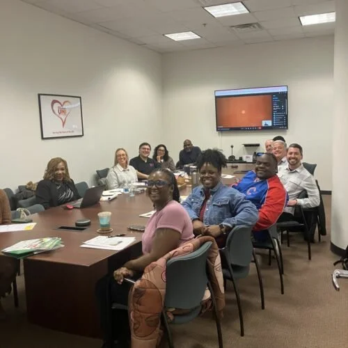 A diverse group of nine people seated around a conference table in a meeting room, smiling and facing the camera, with a large screen mounted on the wall behind them showing a presentation slide or software interface.