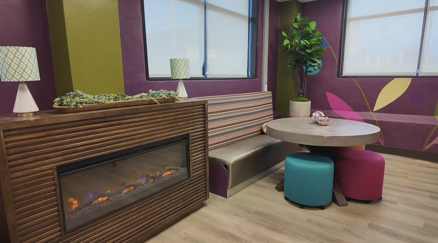 A cozy corner in a room with colorful decor, featuring a fireplace, a bench with striped upholstery, and a round table with two vibrant ottomans, a large potted plant, and purple and green walls decorated with abstract leaf designs.