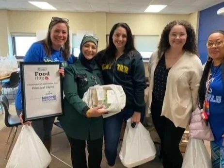 Group of five women standing inside a room, smiling, holding large bags of food and a framed food drive poster.