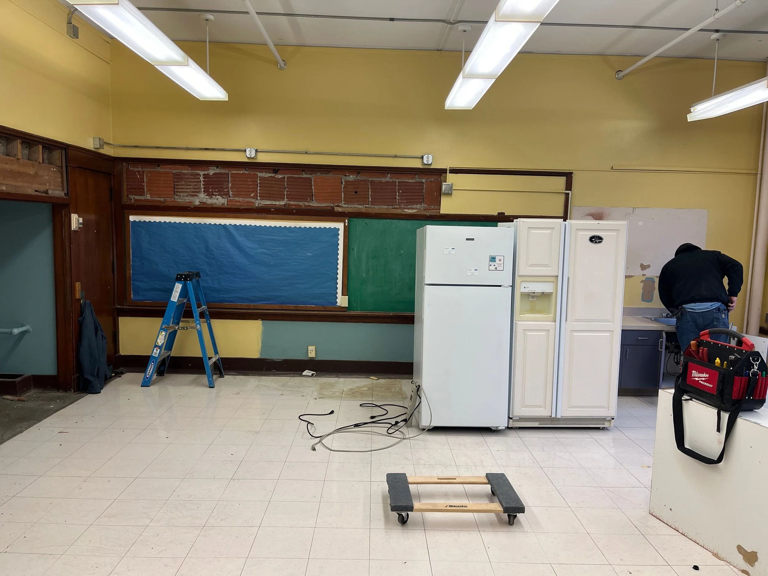 An empty classroom or workshop space with yellow walls, white tiled floor, and ceiling lights. A partially removed wall shows exposed red brick and a large blue bulletin board. There is a small white refrigerator, a tall white cabinet, and a man working near a sink. Various tools and equipment are scattered, including a blue ladder and a wheeled cart.