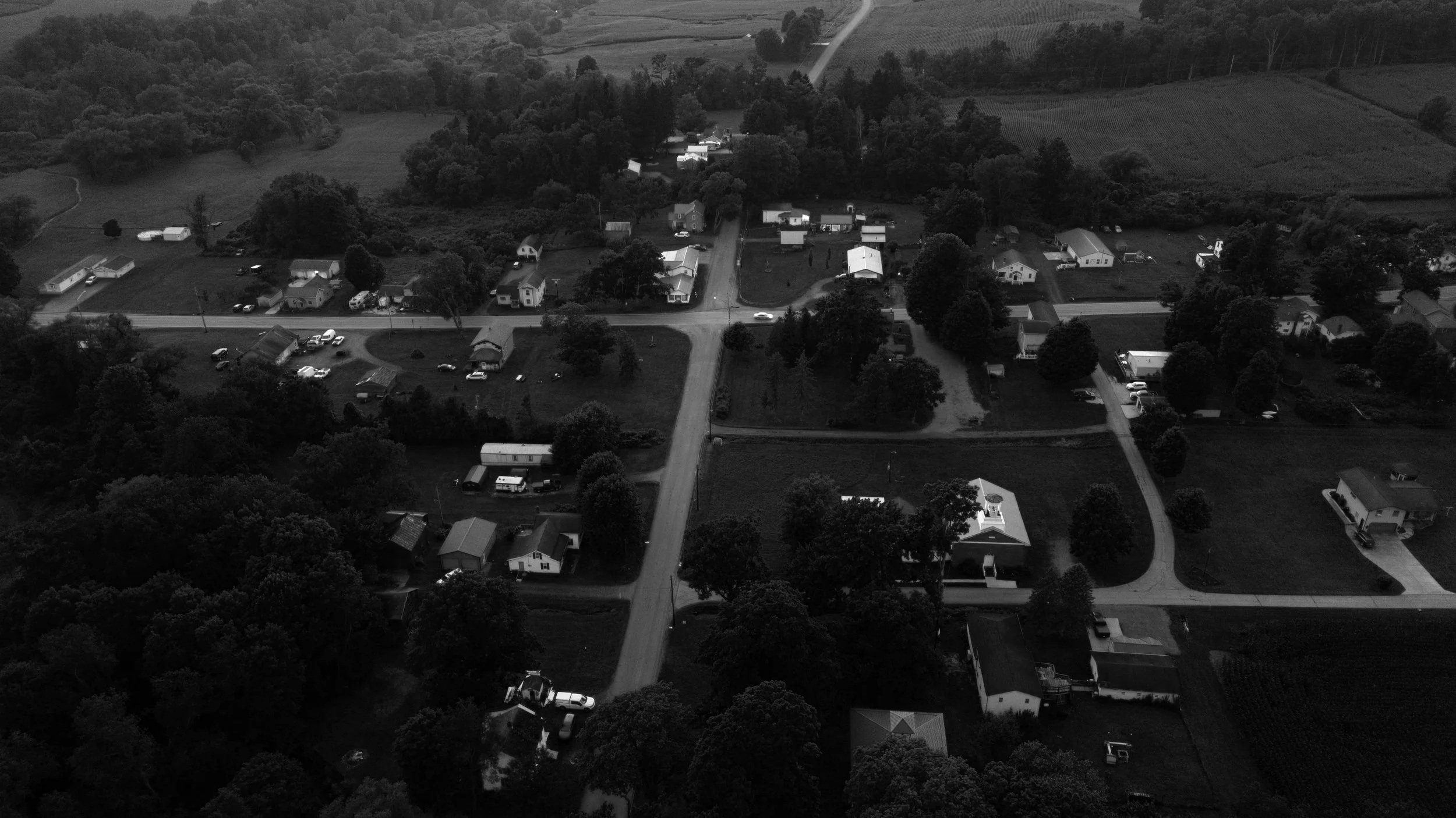 Aerial view of a small rural neighborhood with houses, trees, and roads in a green landscape.