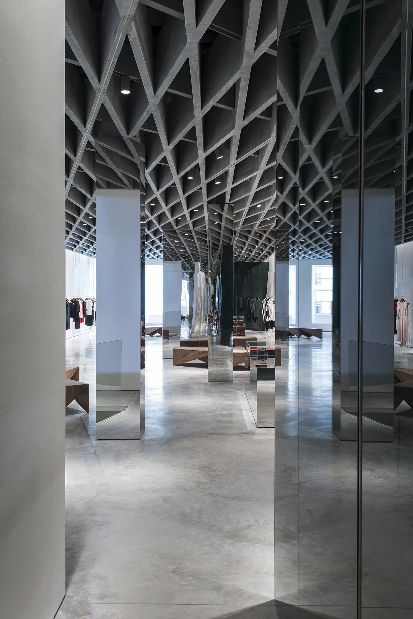 Modern store interior with geometric ceiling design, minimalist displays, and clothing on racks, viewed through glass doors.