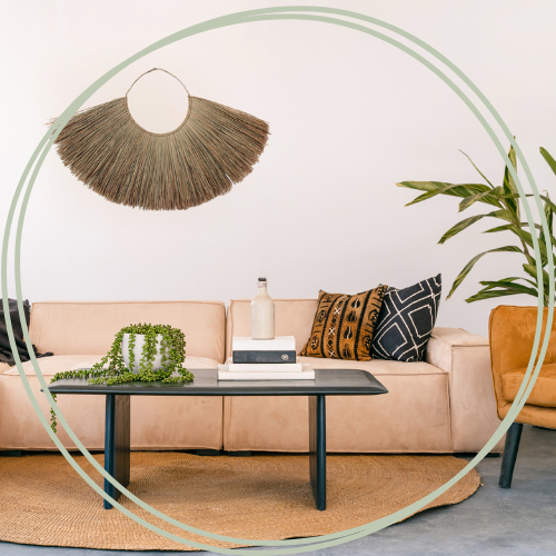 Cozy living room with beige sofa, decorative pillows, a small black coffee table, a bottle on the table, a green plant, and a woven wall hanging.