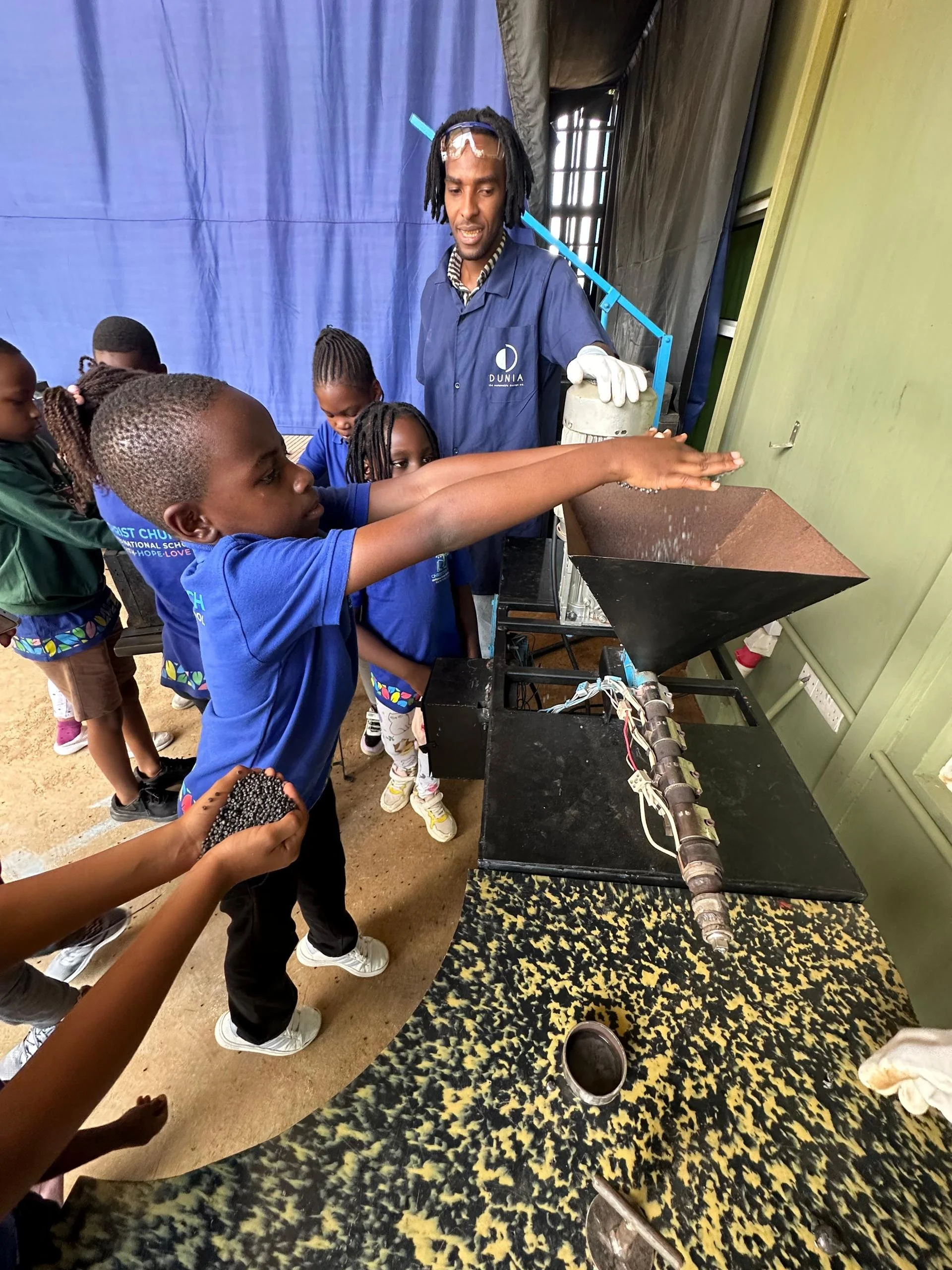 A group of children standing in line, with one child reaching out to touch or operate a machine, while an adult male in a blue shirt observes. The scene appears to be indoors, possibly in a school or educational setting.