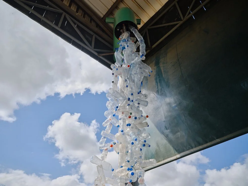 A green funnel shaped object mounted under a roof, pouring out a cascade of plastic bottles into a large basin below.