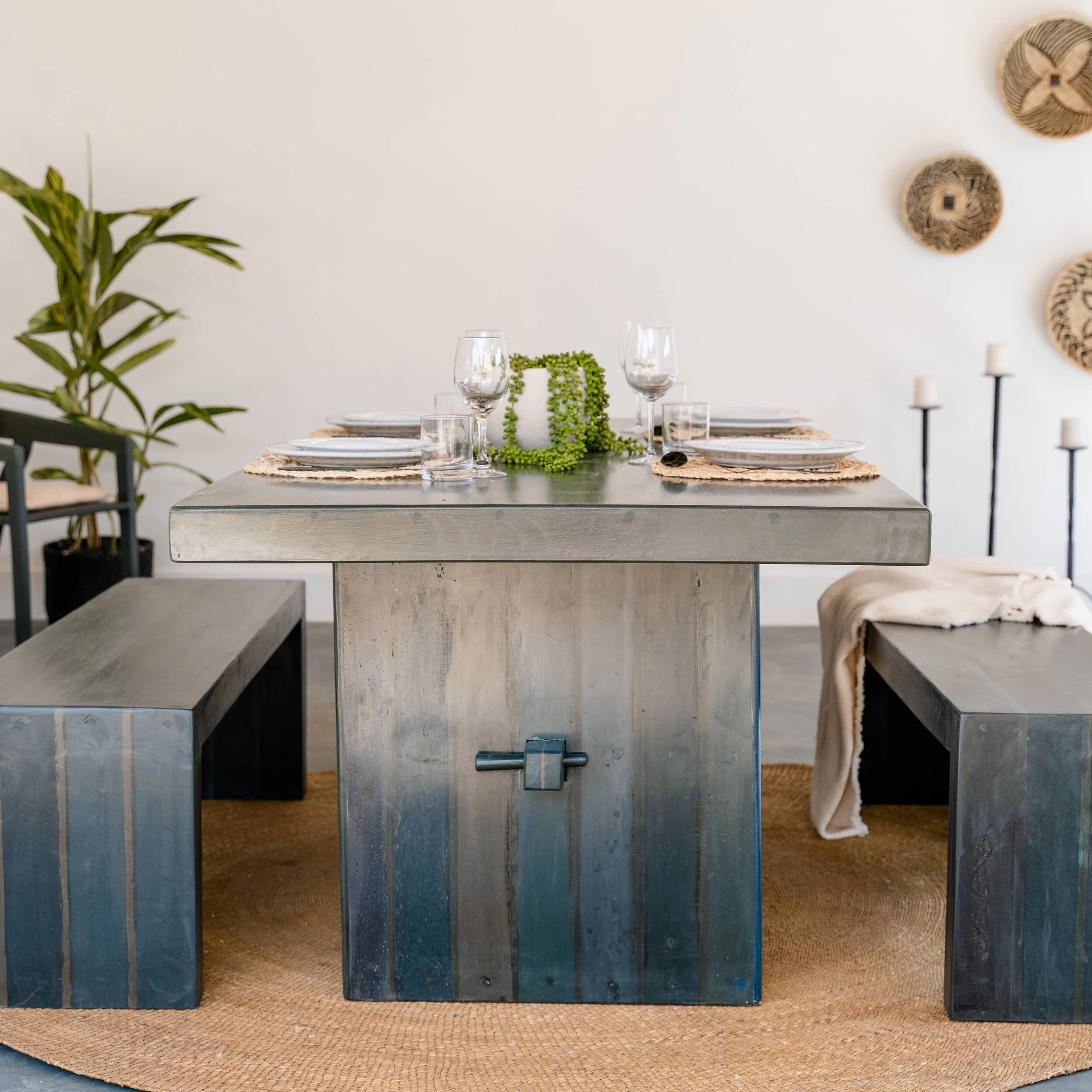 Dining table set with four glasses, plates, a centerpiece, and a woven placemat on a exposed concrete table in a modern dining room. Decor includes woven wall art and a potted plant on the left, with candles on stands on the right.