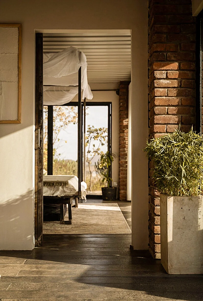 Interior view of a cozy room with a bed covered in white sheets, a large window showing trees outside, exposed brick wall, potted plant, and a wooden floor.