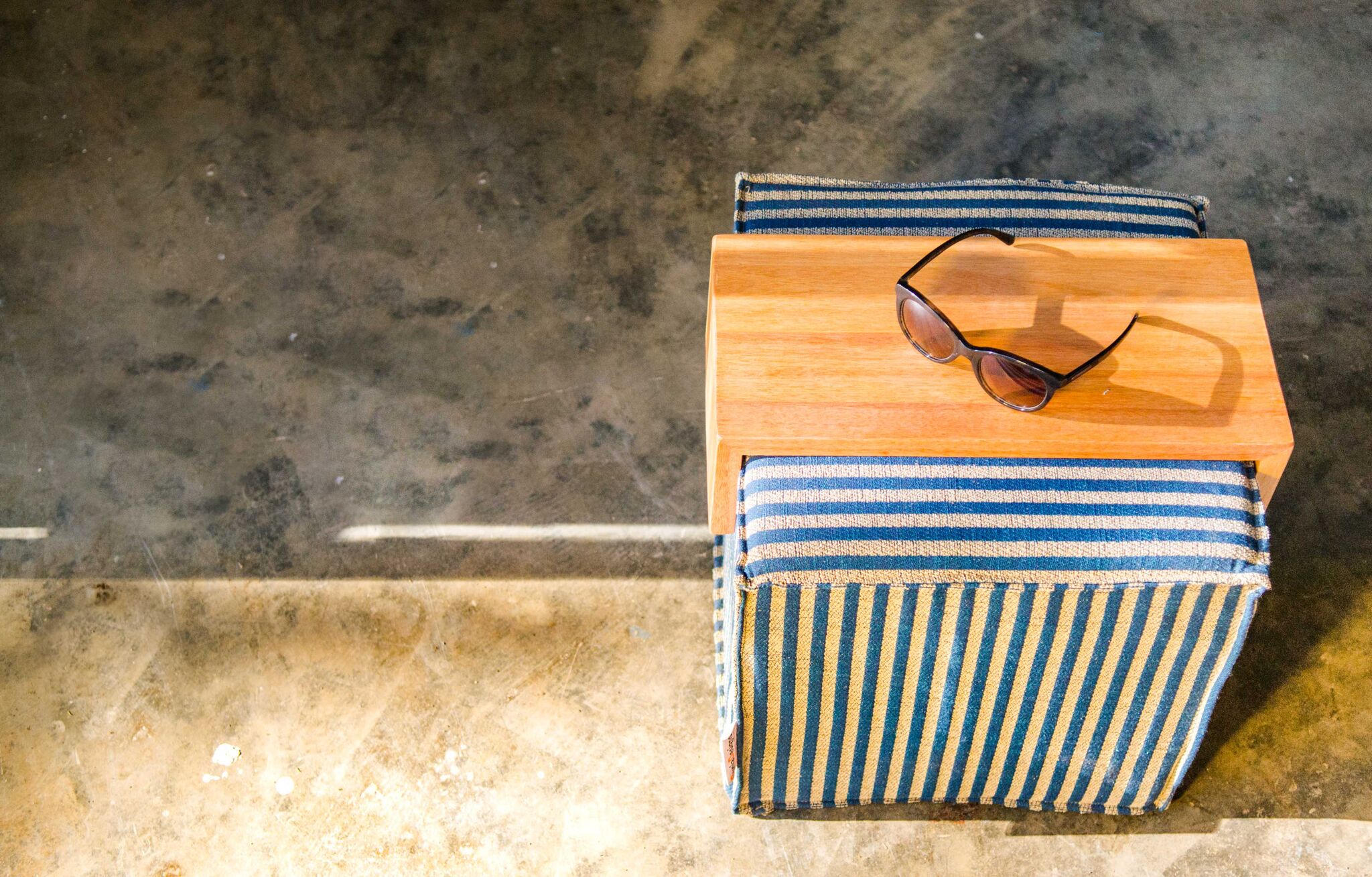 Top view of a small wooden table with a pair of sunglasses on it, placed on a striped cushioned chair against a concrete floor.