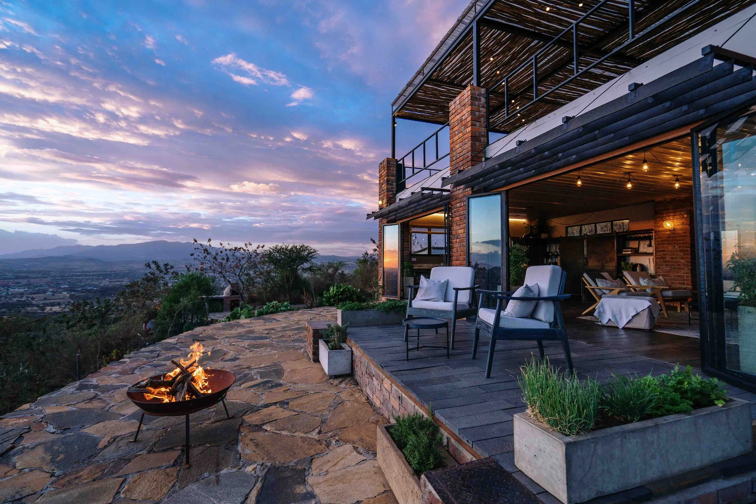 Outdoor terrace with seating area, fire pit, and view of sunset over mountains and sky with clouds.