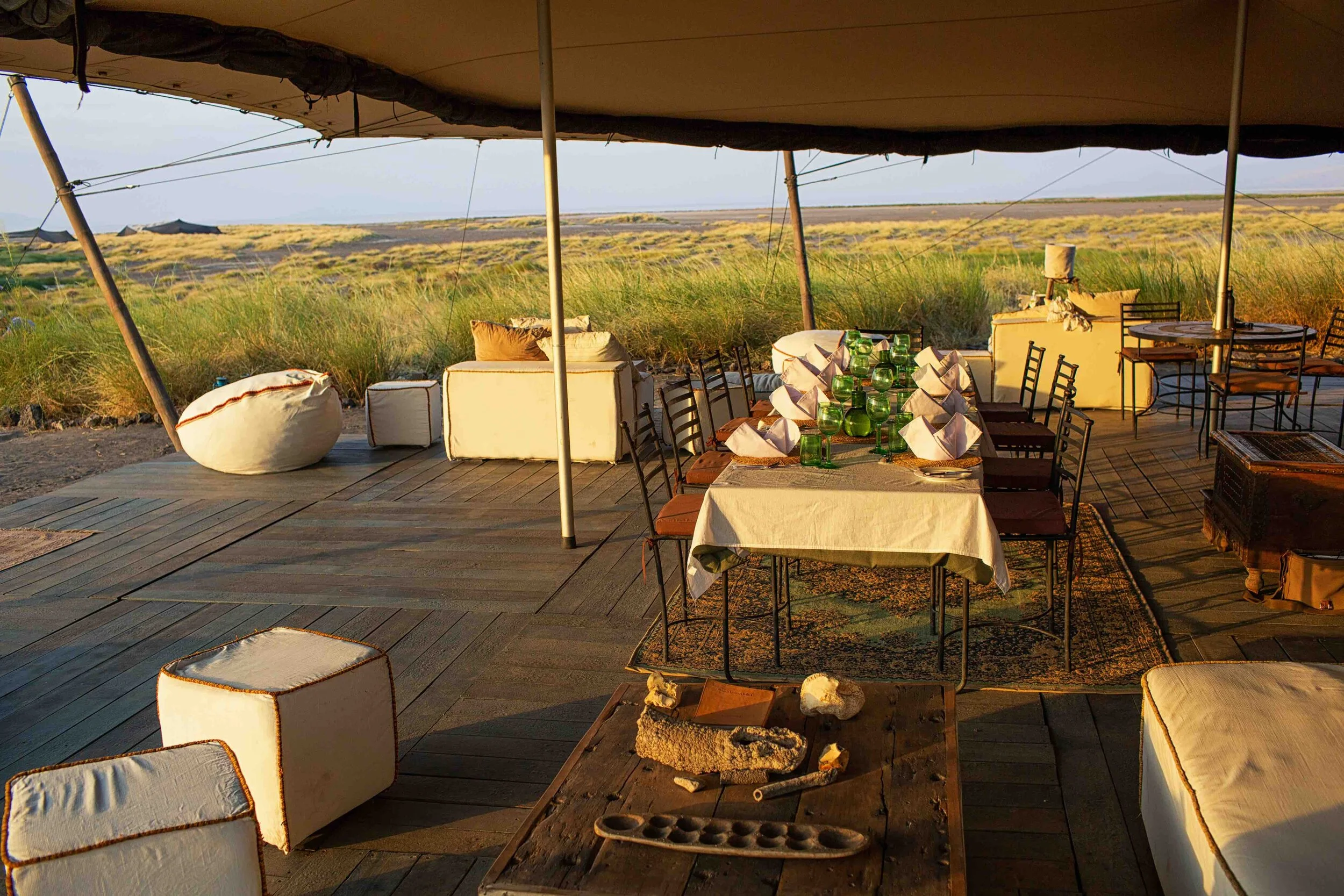 Outdoor dining area with a long table set with white napkins and green glasses, surrounded by chairs and cushioned seats, under a large canopy, with a grassy landscape in the background during sunset.