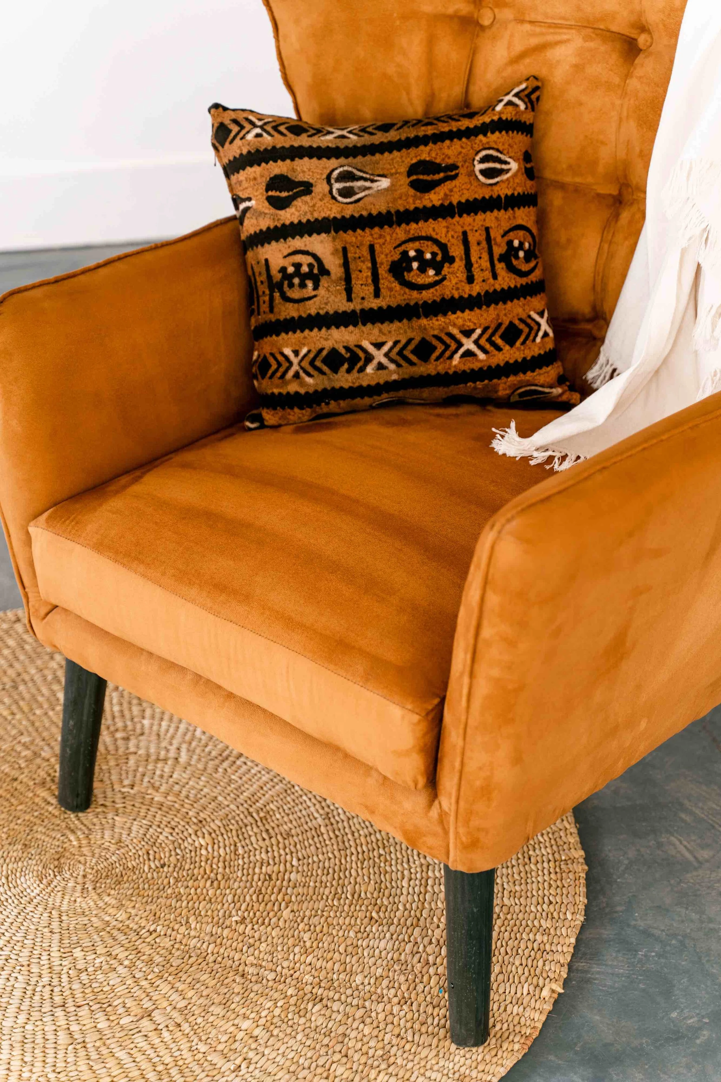 A tan upholstered armchair with black wooden legs, placed on a woven circular rug. There is a decorative pillow with black, white, and tan tribal patterns resting on the chair, and a white fringed throw blanket hanging on the armrest.