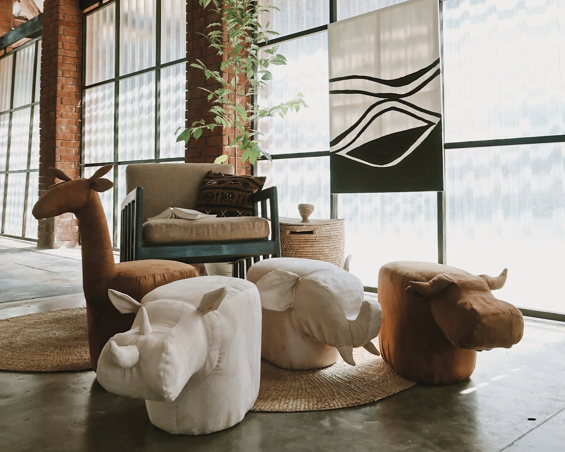 Interior of a rustic industrial-style living space with large windows, exposed brick walls, and a mix of furniture including a stuffed animal resembling a horse, a beige armchair, woven side table, and assorted ottomans and cushions, accented by a tall plant and modern wall art.