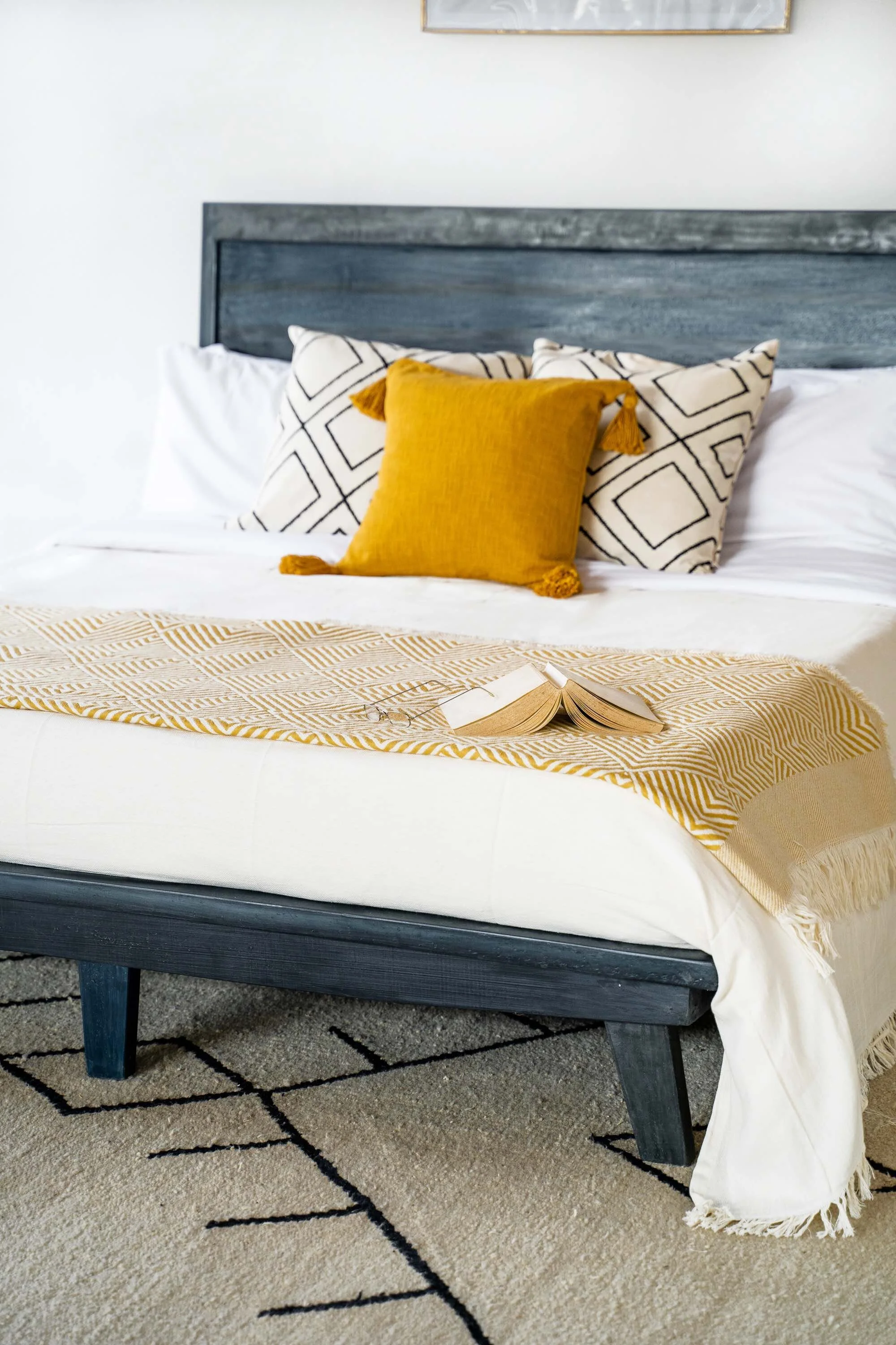 A bed with a white duvet and a beige and white patterned throw blanket, topped with a yellow pillow, and two white pillows with black geometric patterns. An open book is on the bed, and a fringed white throw is hanging over the edge. The bed has a gray wooden headboard, and a framed picture is partly visible on the white wall above.