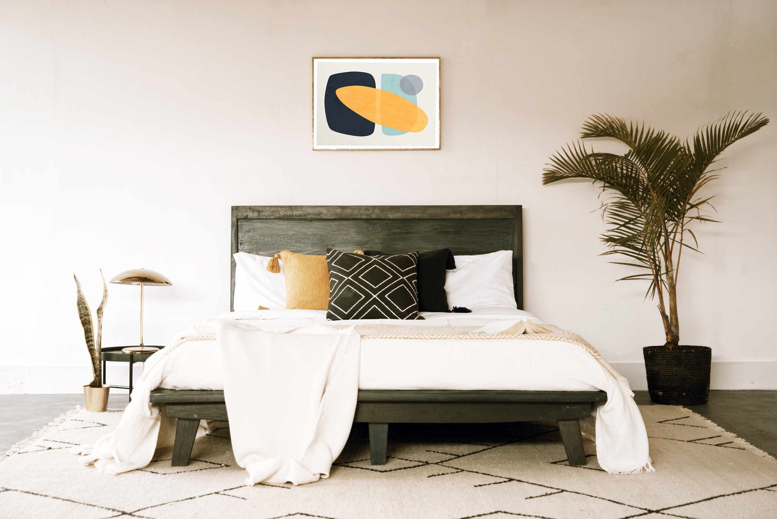 A bedroom with a wooden bed, white bedding with black, yellow, and beige pillows, a patterned rug, a side table with a lamp, large palm plant in a basket, and abstract artwork on the plain white wall.