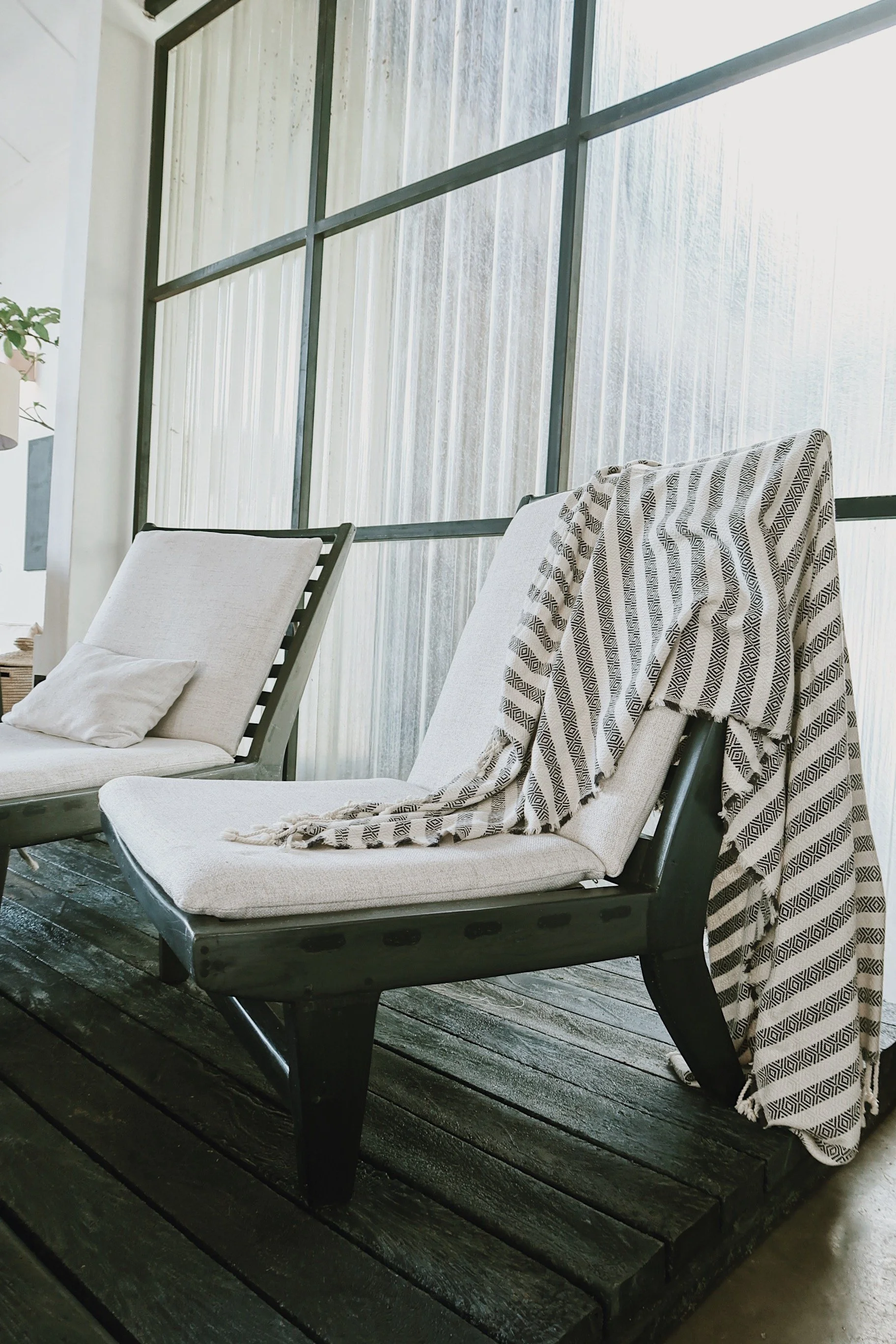 Two lounge chairs with beige cushions and pillows on a dark wooden deck next to a large window. One chair has a patterned towel draped over it.