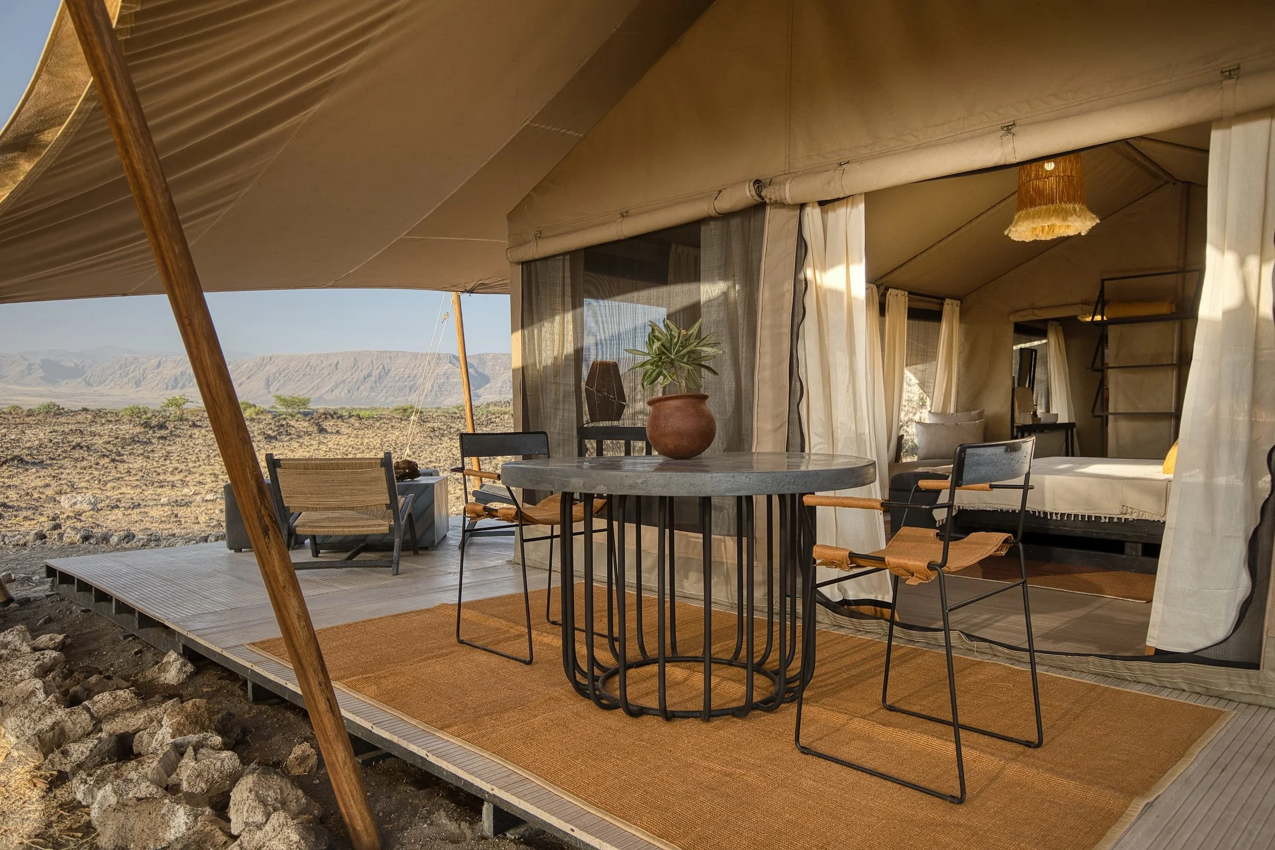 Outdoor camping tent with a patio-style deck, black metal table with a potted plant, surrounding chairs, and a cozy interior visible through the open curtains, set against a desert landscape with mountains in the background.