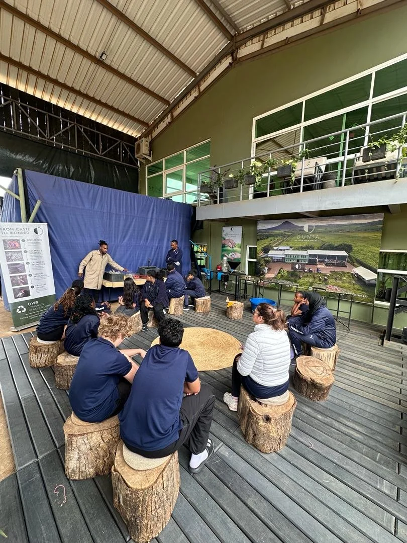 Group of people seated on wooden stumps in an indoor space with a stage, two standing presenters, and a large poster in the background.