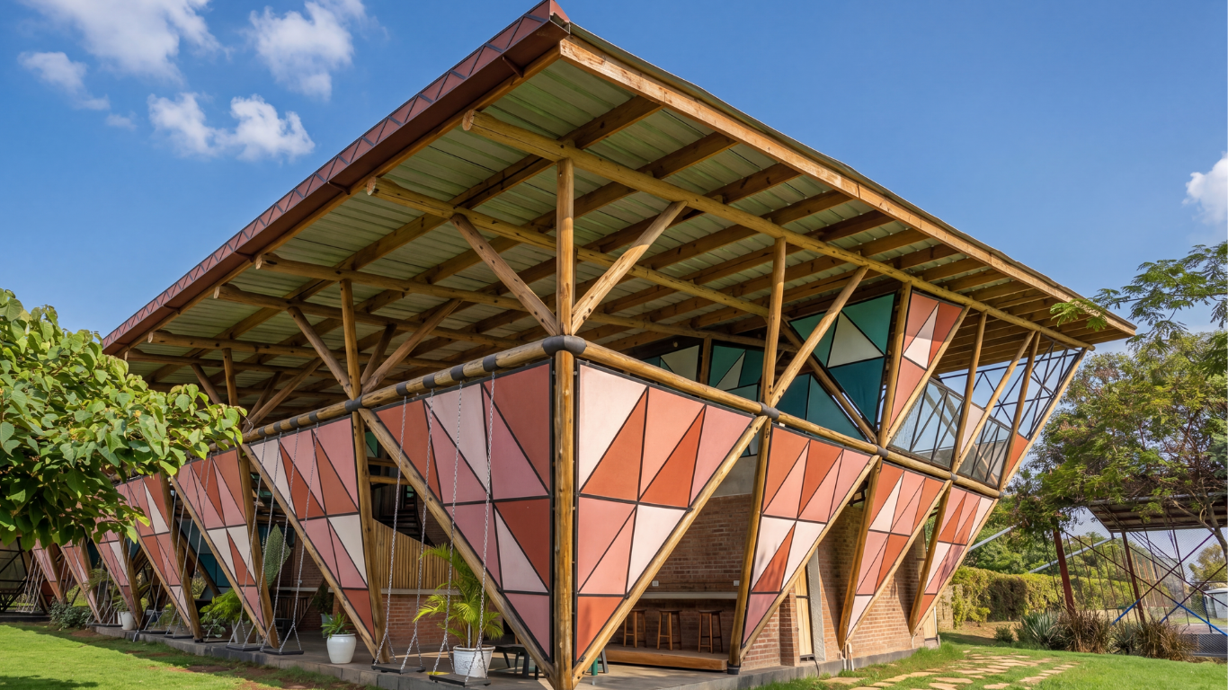 Modern building with geometric pink and teal panels and wooden beams, surrounded by greenery and blue sky.