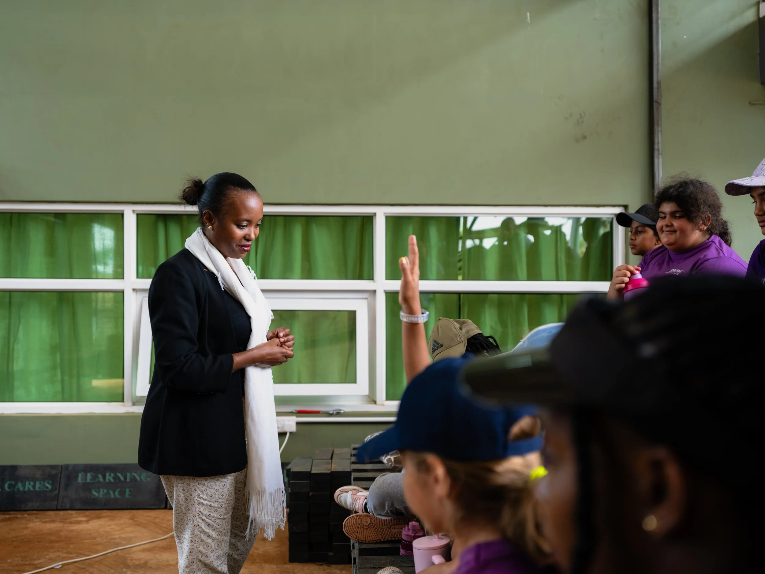 A woman with dark hair tied back, wearing a black blazer and white scarf, stands in front of children raising their hands in a classroom or workshop setting with a green wall and window behind her.