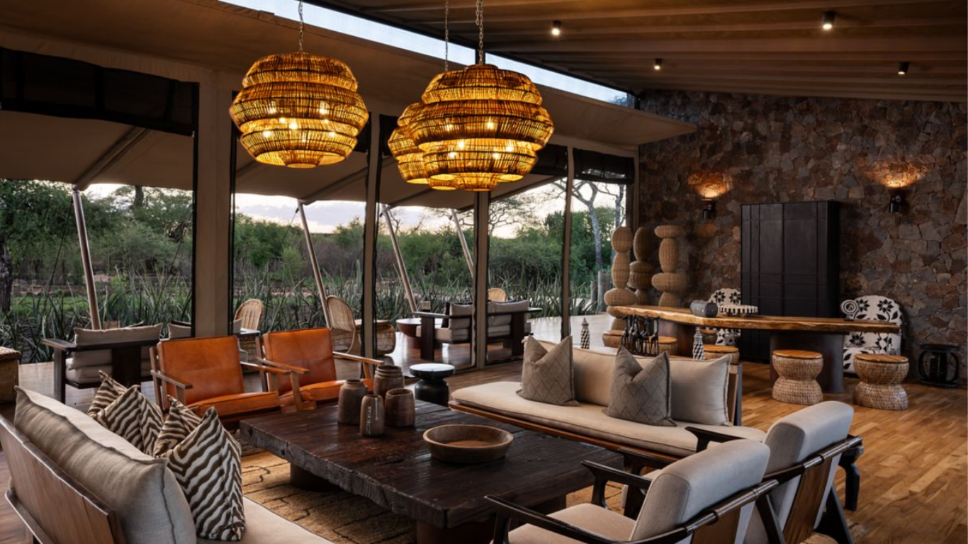 Interior of a modern living room with hanging wicker pendant lights, wooden furniture, and large glass sliding doors showing an outdoor view of trees and greenery.