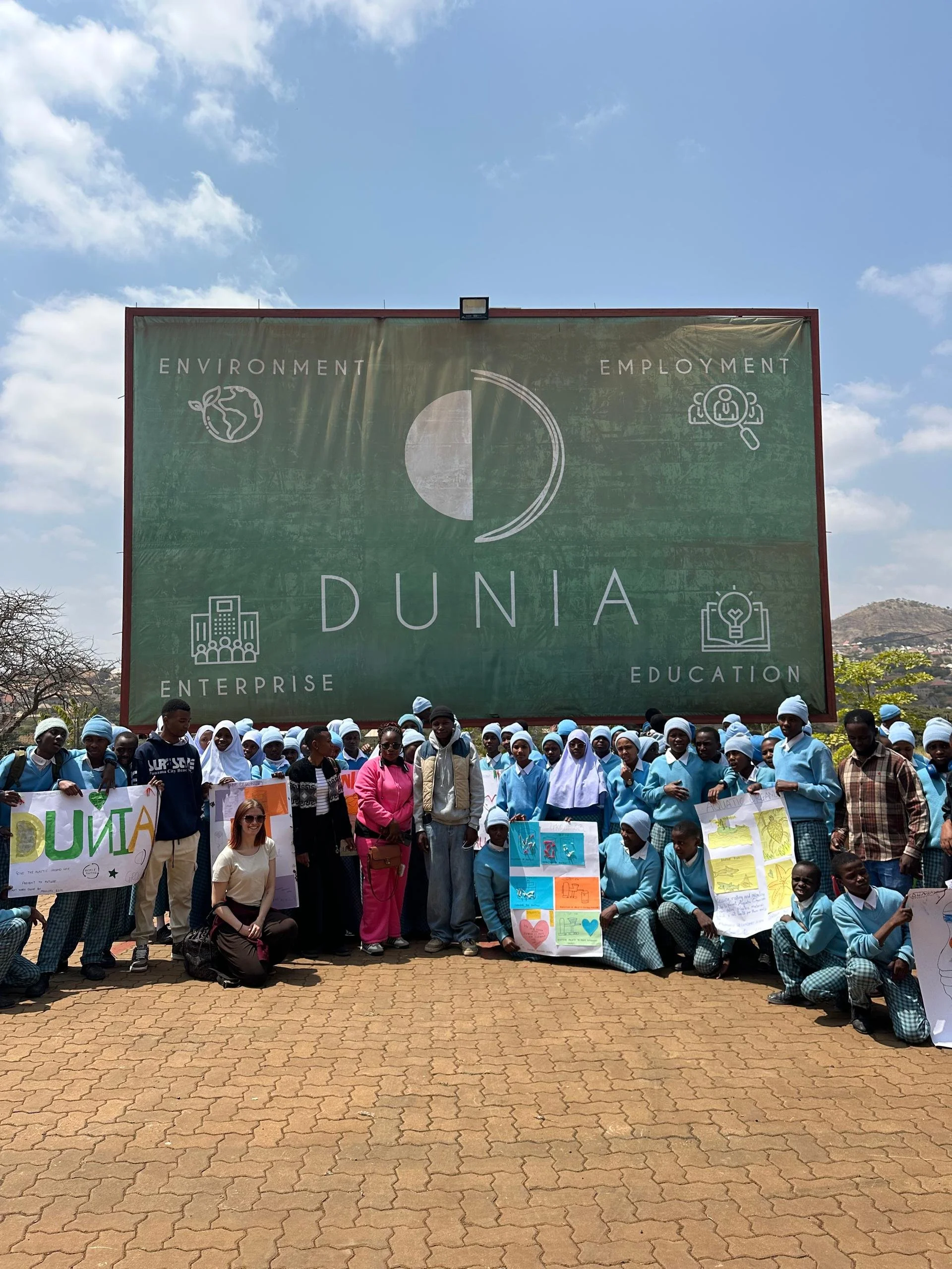 A large group of students and adults holding posters and standing in front of a big green billboard with the words "Dunia" and icons representing environment, employment, enterprise, and education.