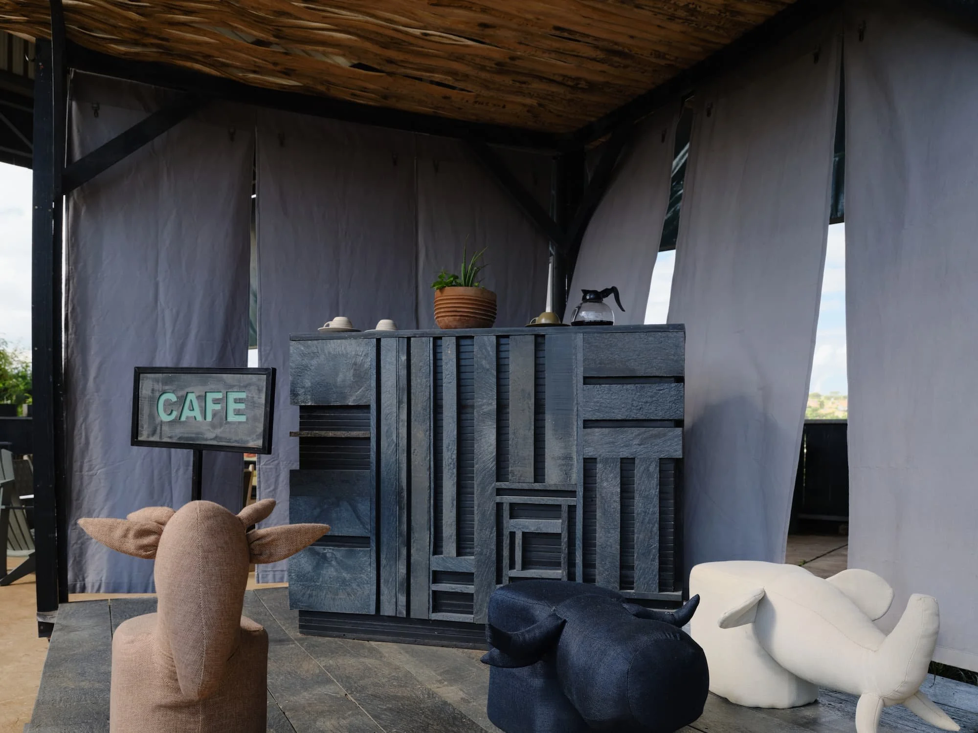 Outdoor cafe setup with a gray wooden counter, potted plant, cups, a glass pitcher, and decorative animal figures in the foreground, and a gray curtain backdrop with a wooden roof overhead.