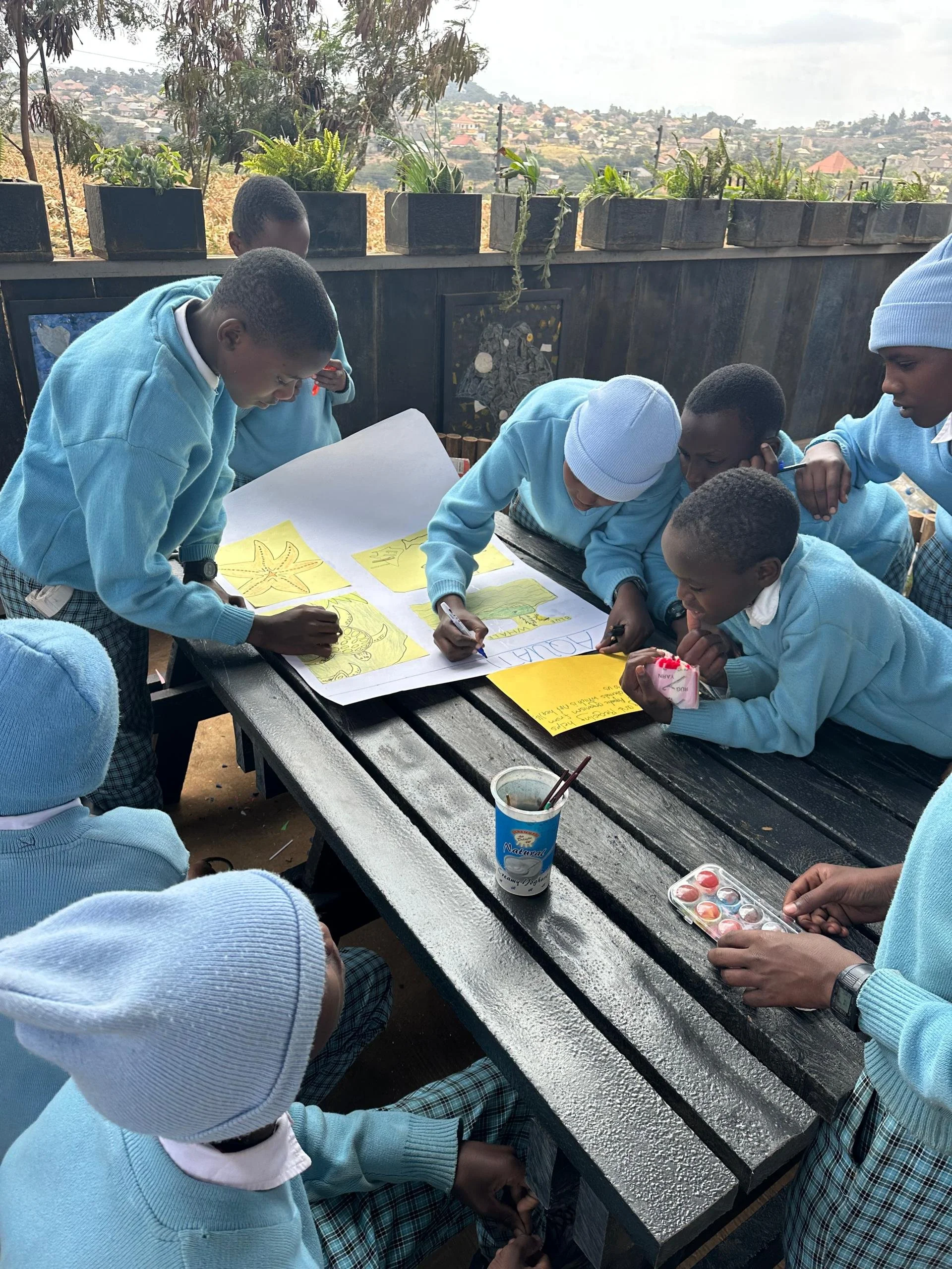 Group of children and teenagers wearing light blue sweaters and plaid trousers, gathered around a dark wooden table outdoors, engaging in drawing and painting activities with paints, brushes, and paper, with a scenic view of houses and trees in the b