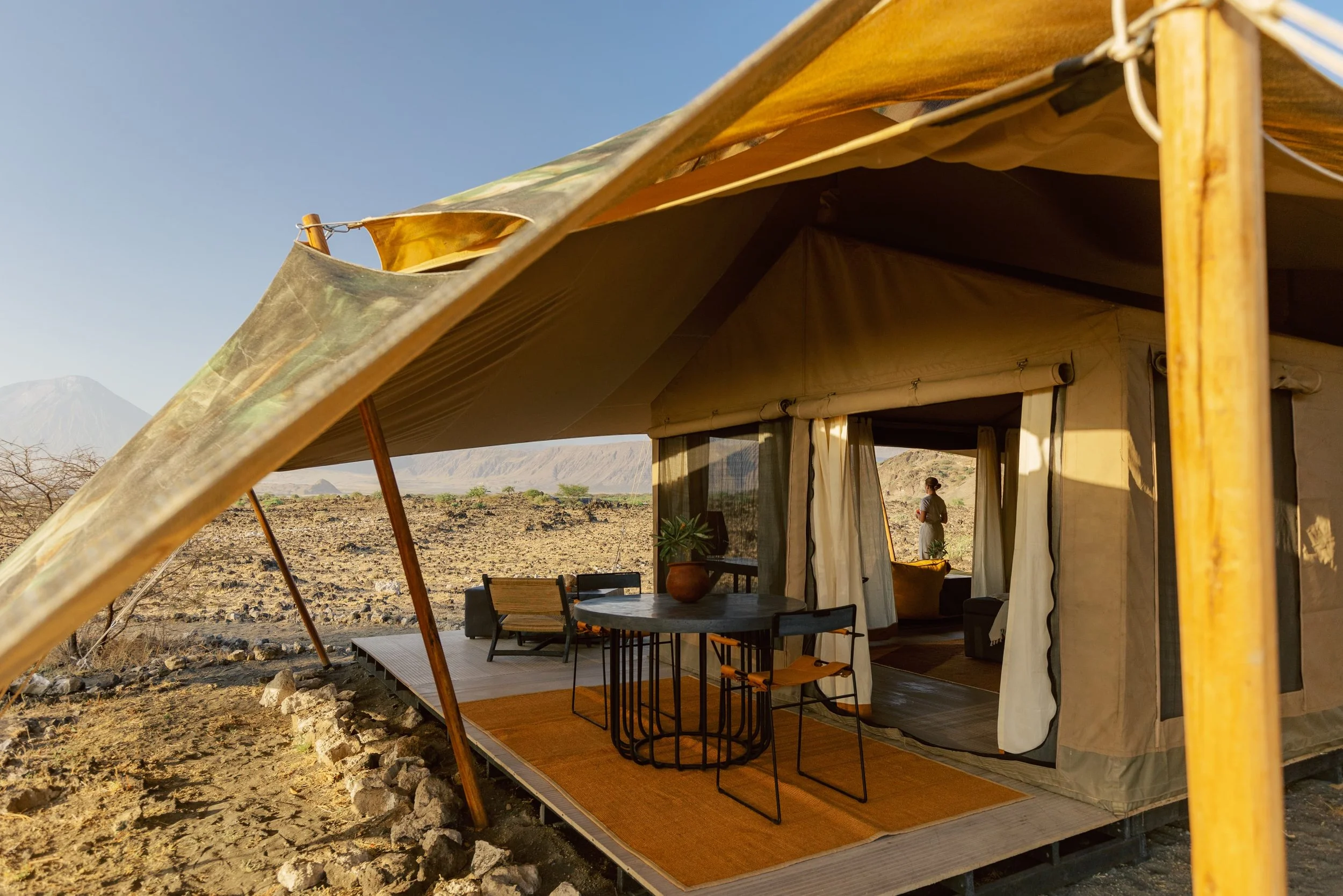 View of a desert landscape with a tented outdoor living area featuring a wooden table, chairs, and a potted plant under a canopy near Mount Kilimanjaro in the background.