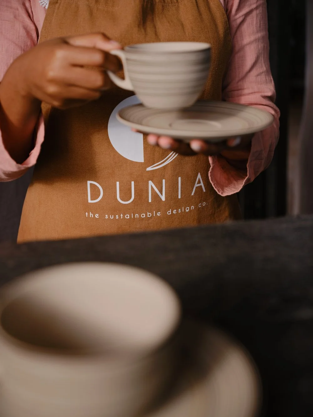Person holding a ceramic cup and saucer, wearing a brown apron with the word "DUNIA" and environmental message, in a pottery or coffee shop setting.