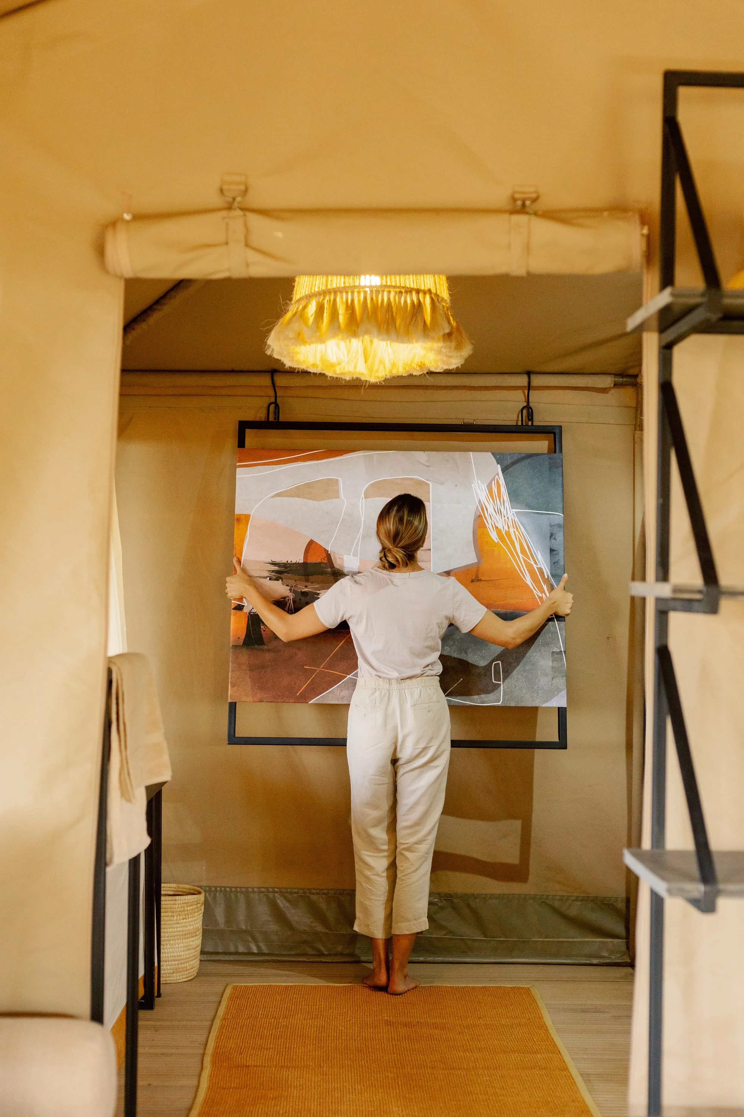 Person standing in front of a large abstract art piece in a cozy, warmly lit room. The individual is seen from behind, wearing a white t-shirt and light-colored pants, with arms extended to hold the artwork. The room features beige walls, a hanging y
