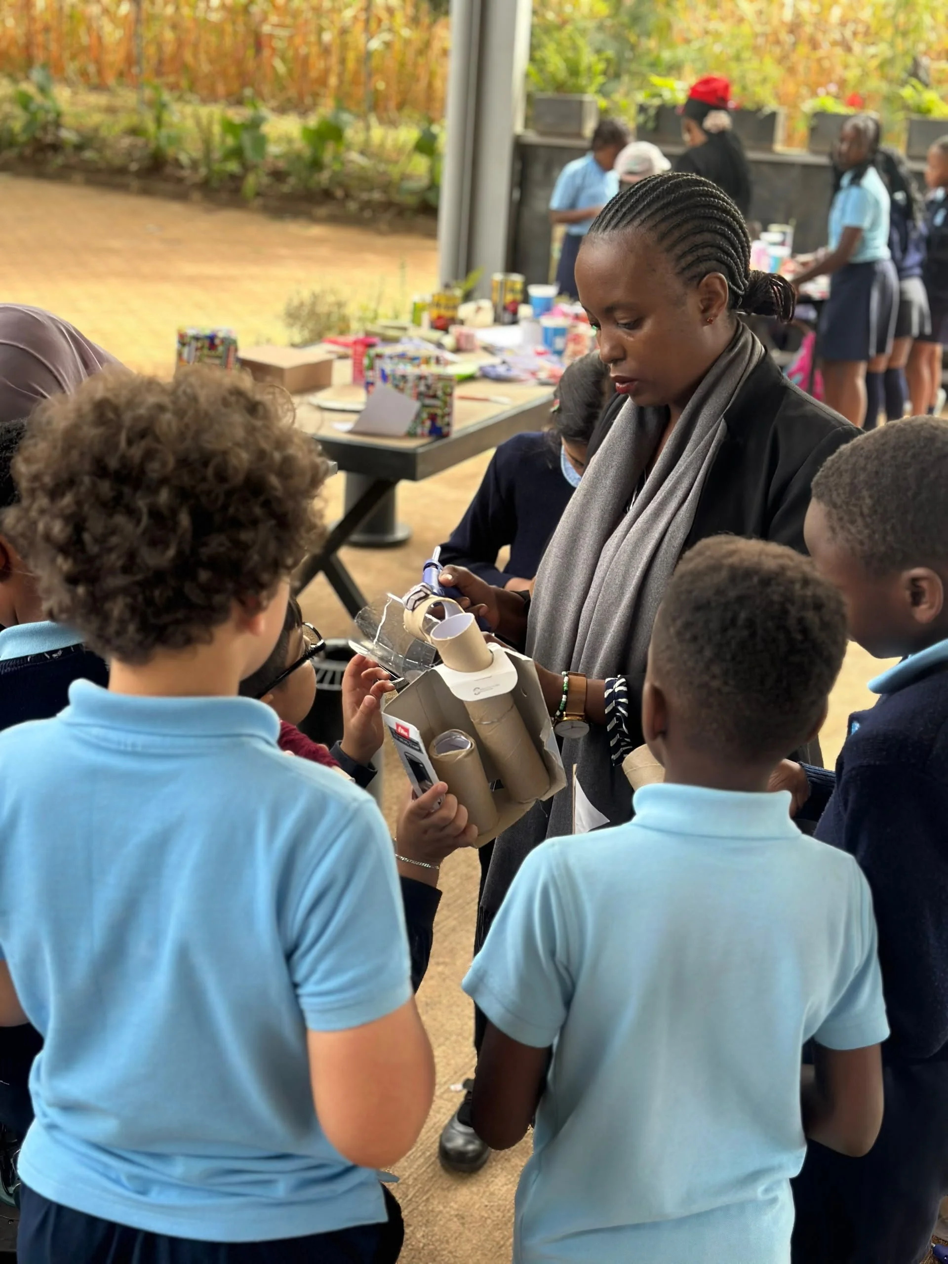Group of children and a woman gathered around a table with school supplies outside. The children are wearing light blue uniforms, and the woman is holding a cardboard box. In the background, there are more children in uniforms and trees with autumn foliage.