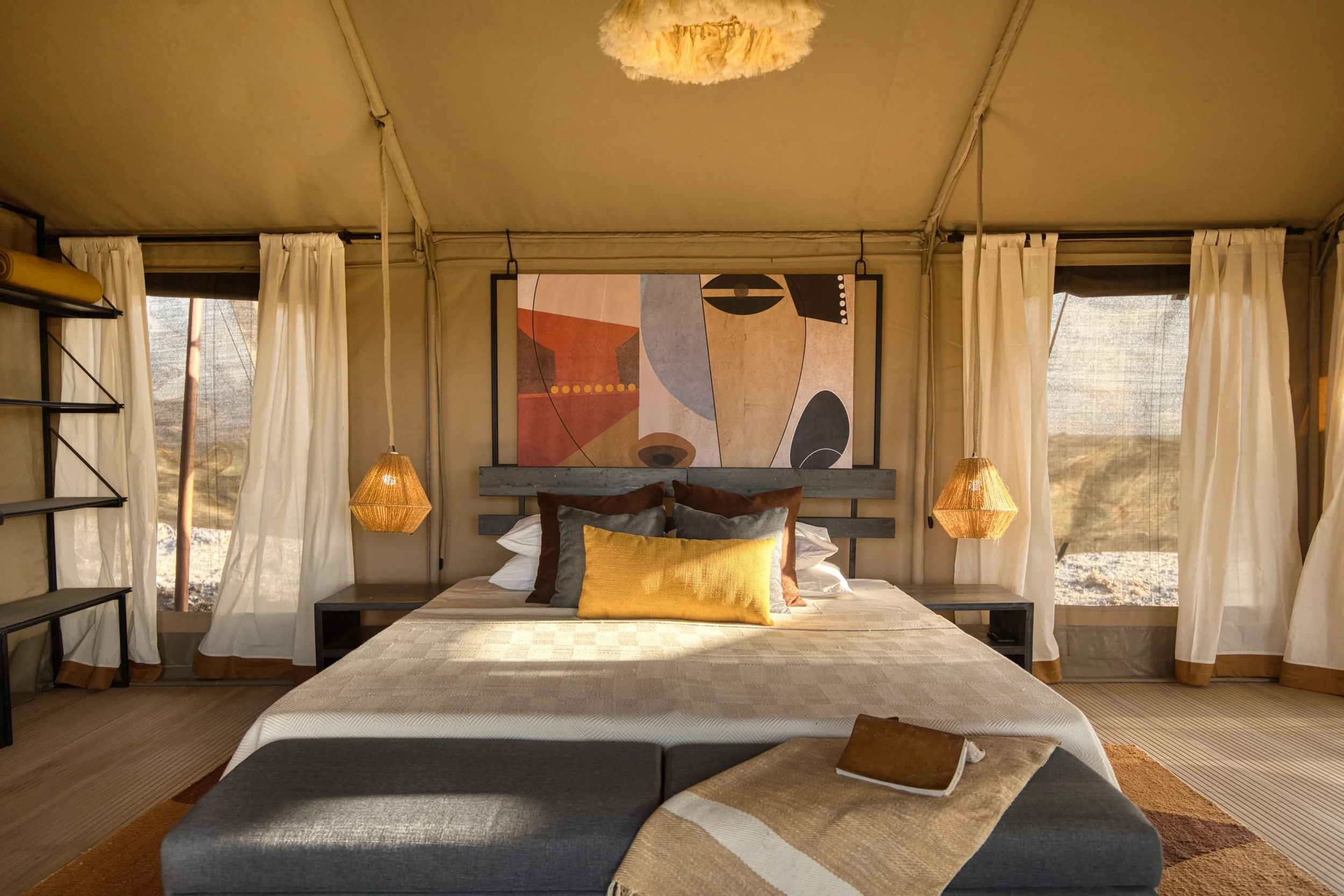 Luxury bedroom with a bed, pillows, and abstract artwork above the headboard, surrounded by curtains and warm lighting in a desert setting.