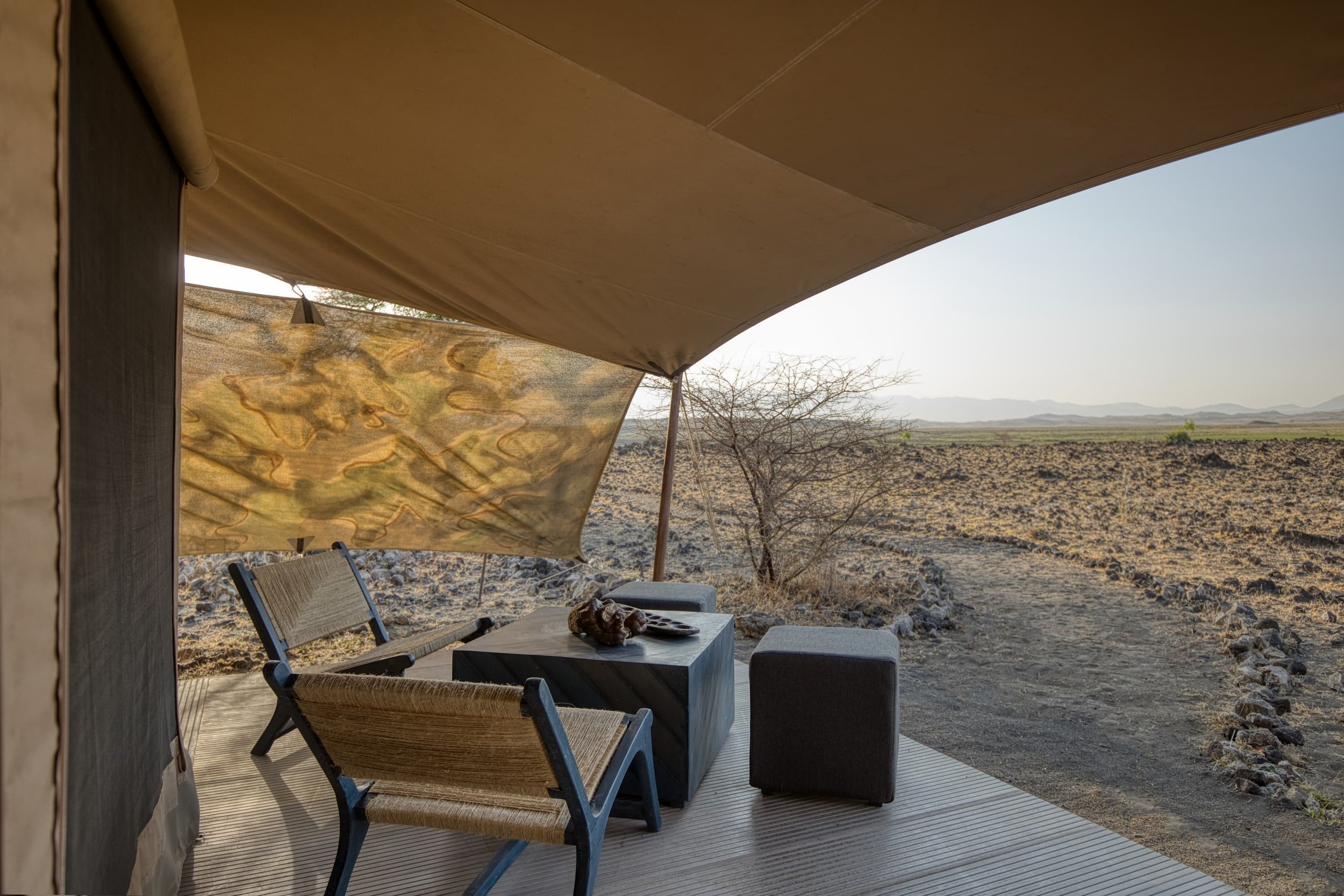Outdoor seating area with two woven chairs facing a small table with decorative objects, surrounded by a desert landscape with rocky ground and sparse bushes, shaded by a large canopy