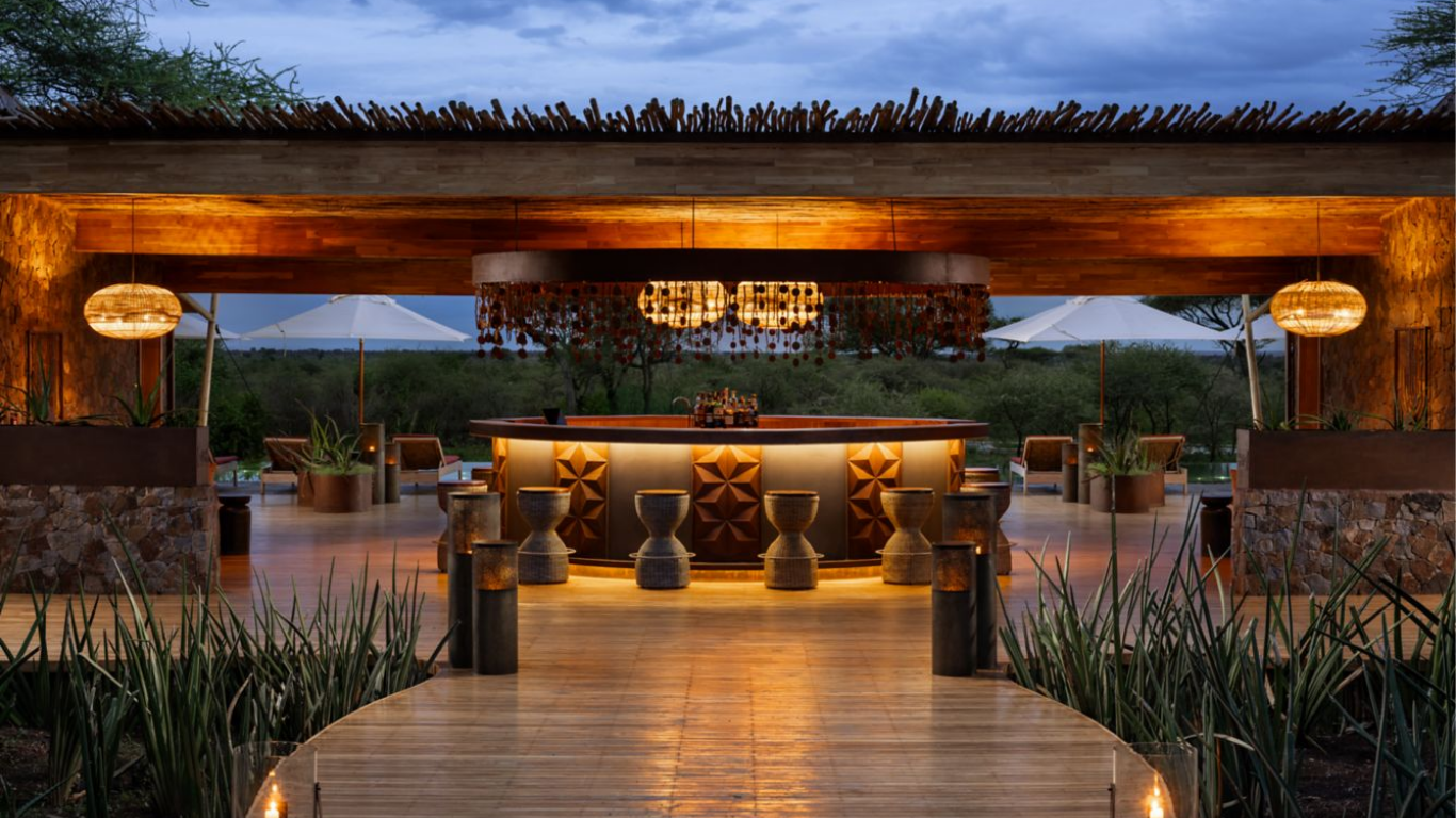 An outdoor bar area with warm lighting, wooden accents, and surrounding greenery, set against a backdrop of trees and a cloudy sky.
