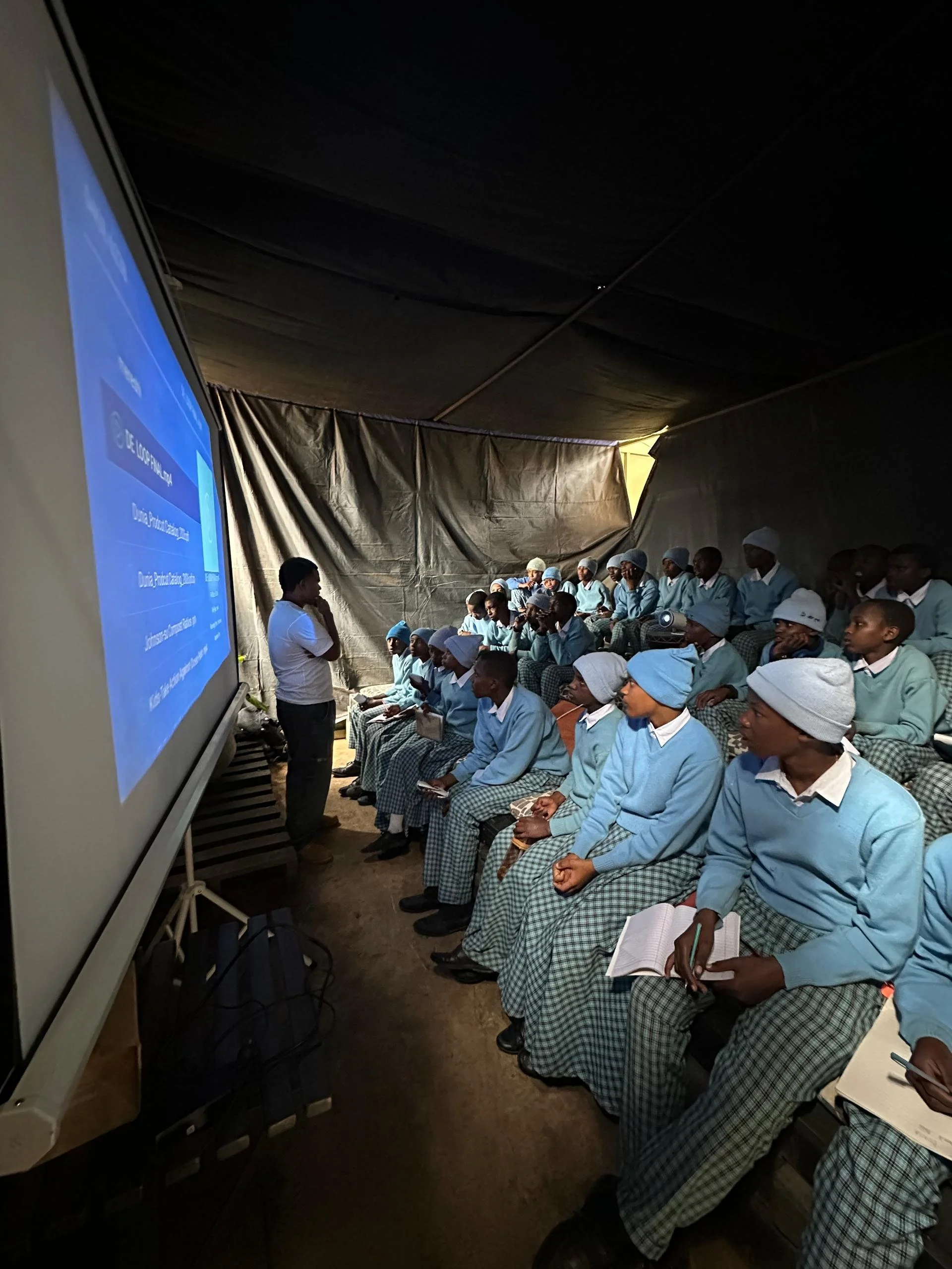 A group of students in school uniform attending a classroom training session, seated and listening to a woman instructor with a projector screen visible.