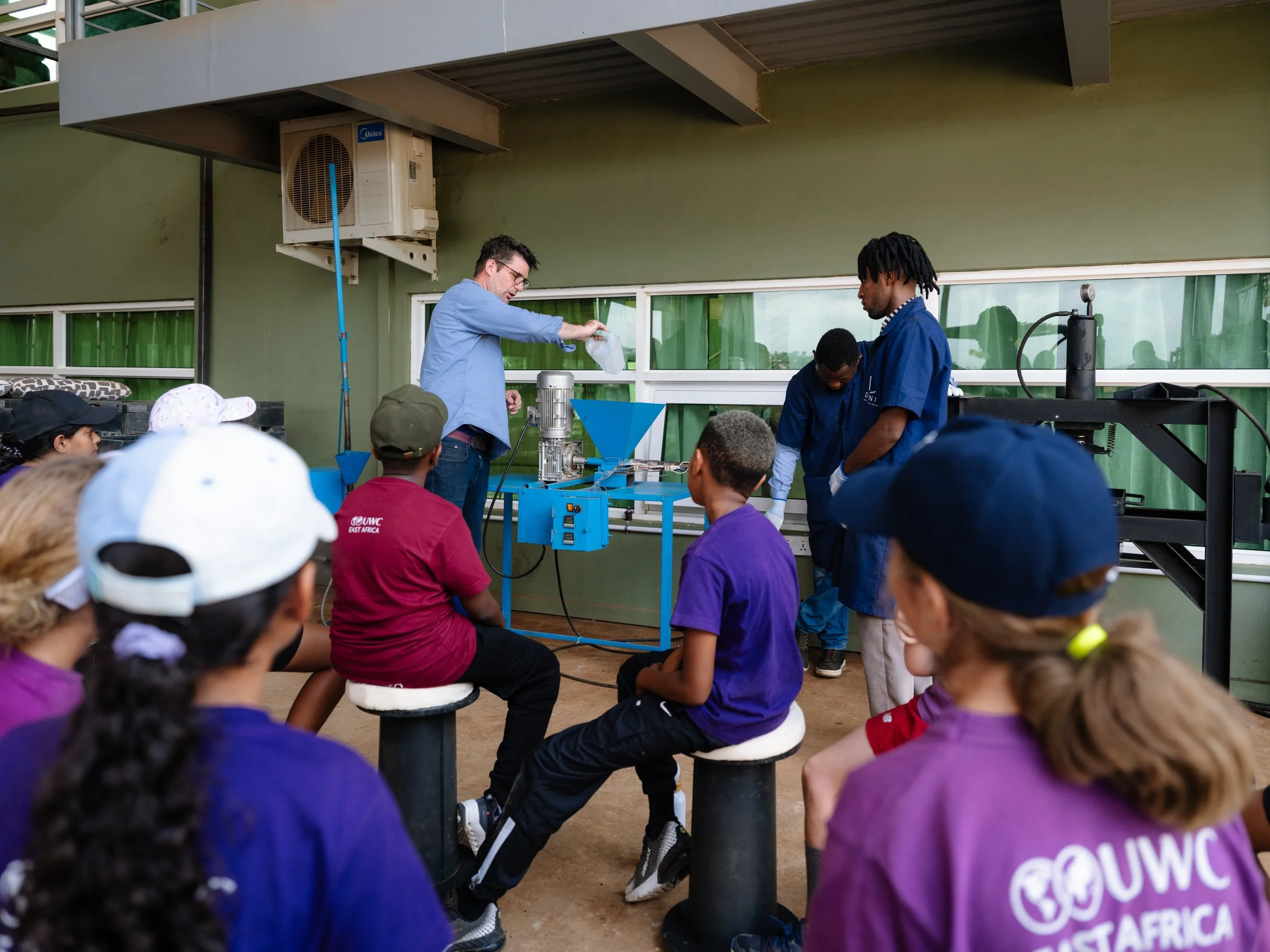A man in a blue shirt conducting a science demonstration in front of a group of children seated on stools. The demonstration involves scientific equipment, and other men in blue shirts are assisting. The children are attentively watching the experime