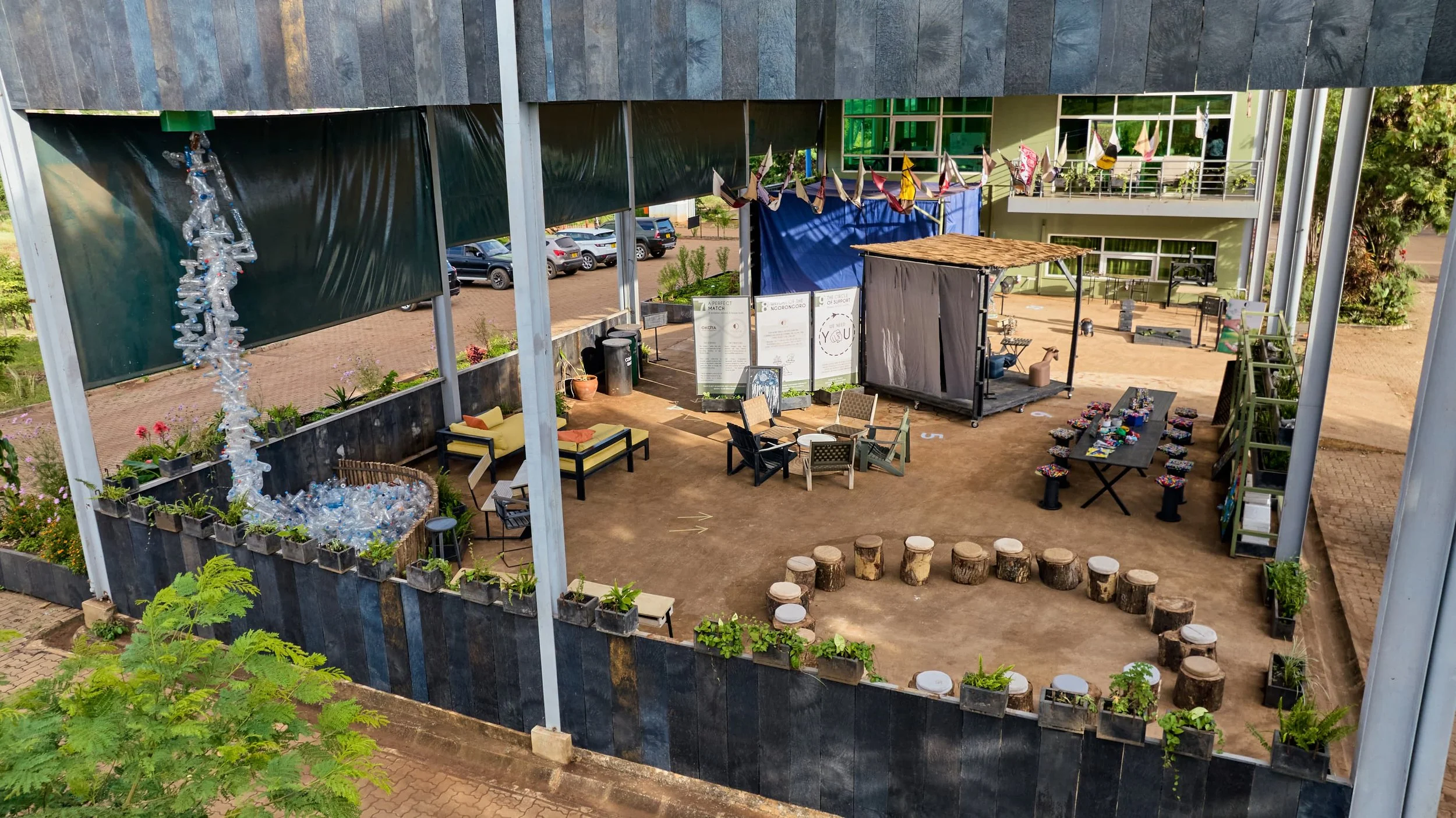 Open-air venue with black walls, string lights, small seating area with chairs and tables, a small stage with a wooden roof, colorful banners, and decorative elements, surrounded by greenery and parked cars in the background.