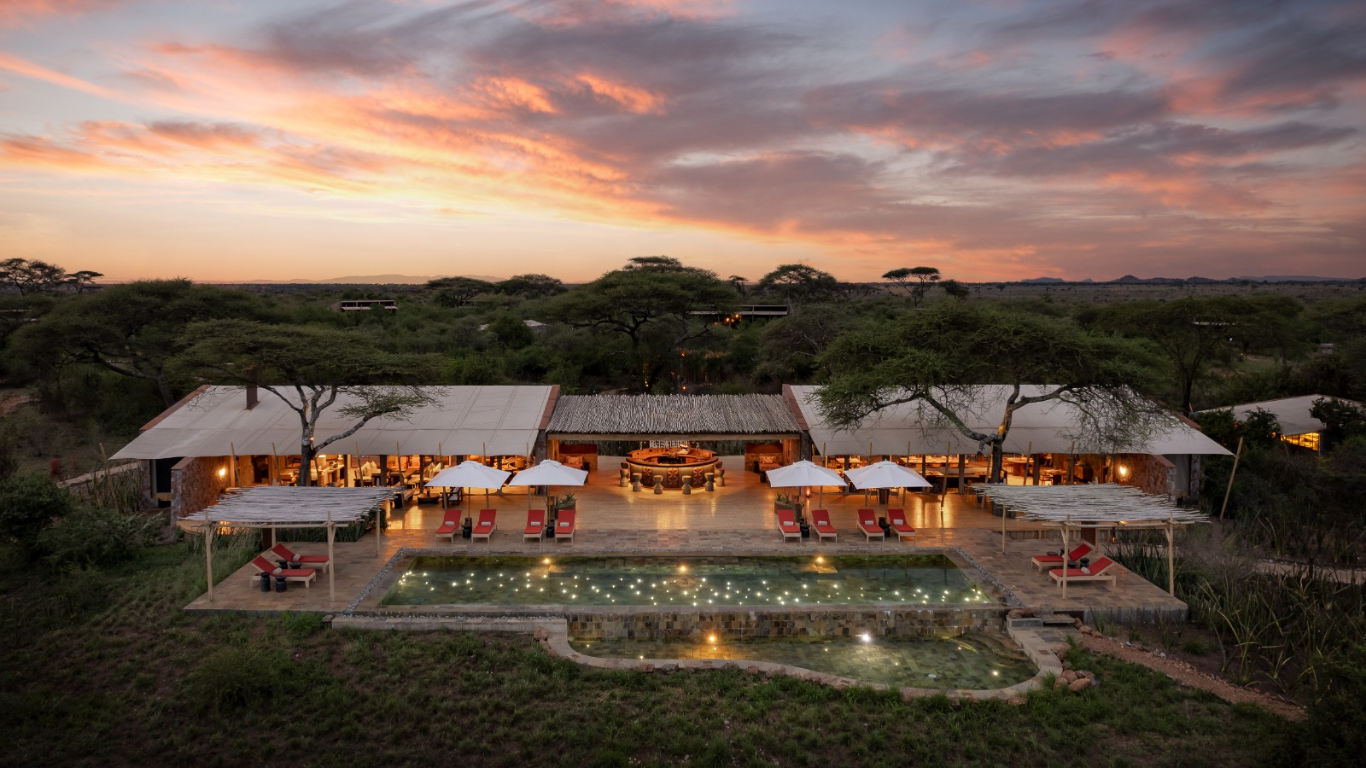 Aerial view of a luxurious outdoor resort in a savanna landscape at sunset. The resort features a large swimming pool with underwater lights, multiple lounge chairs with umbrellas, and a spacious open-air bar and dining area with cozy lighting, surrounded by trees and open plains.