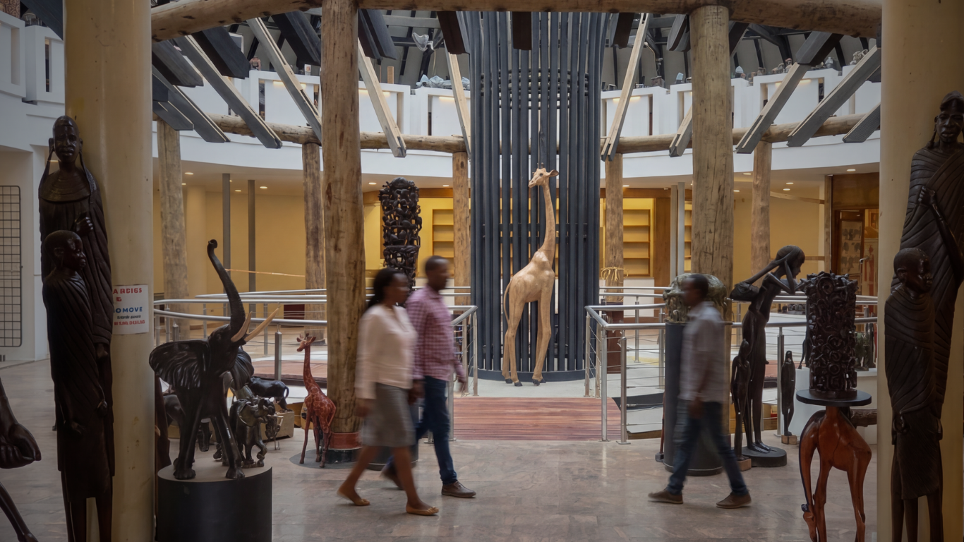 Interior of a museum with sculptures, including a tall giraffe statue and various carved figures, and three visitors walking through.