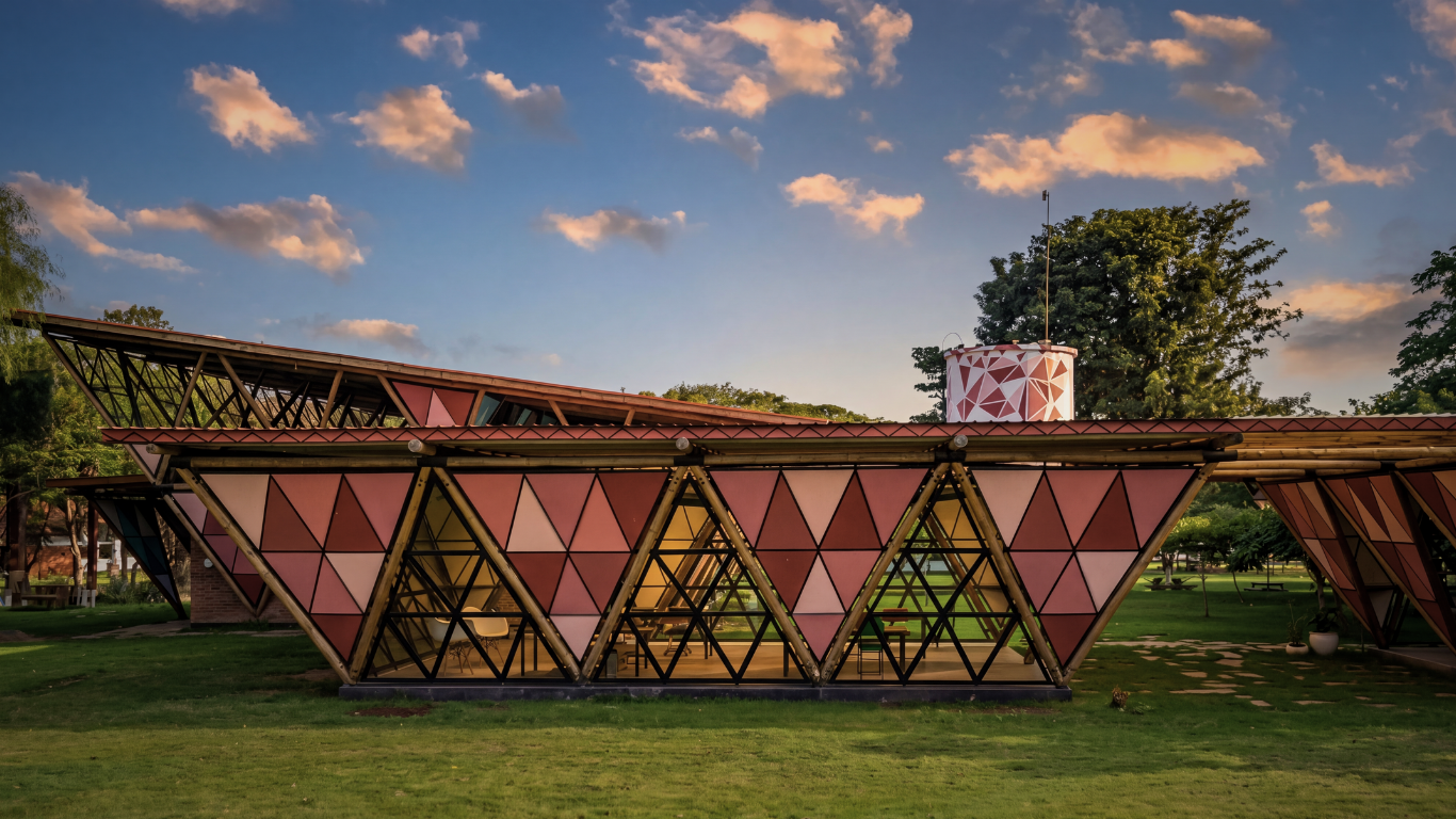Modern geometric building with pink triangular panels and black metal framing, set in a green park during sunset with a partly cloudy sky.