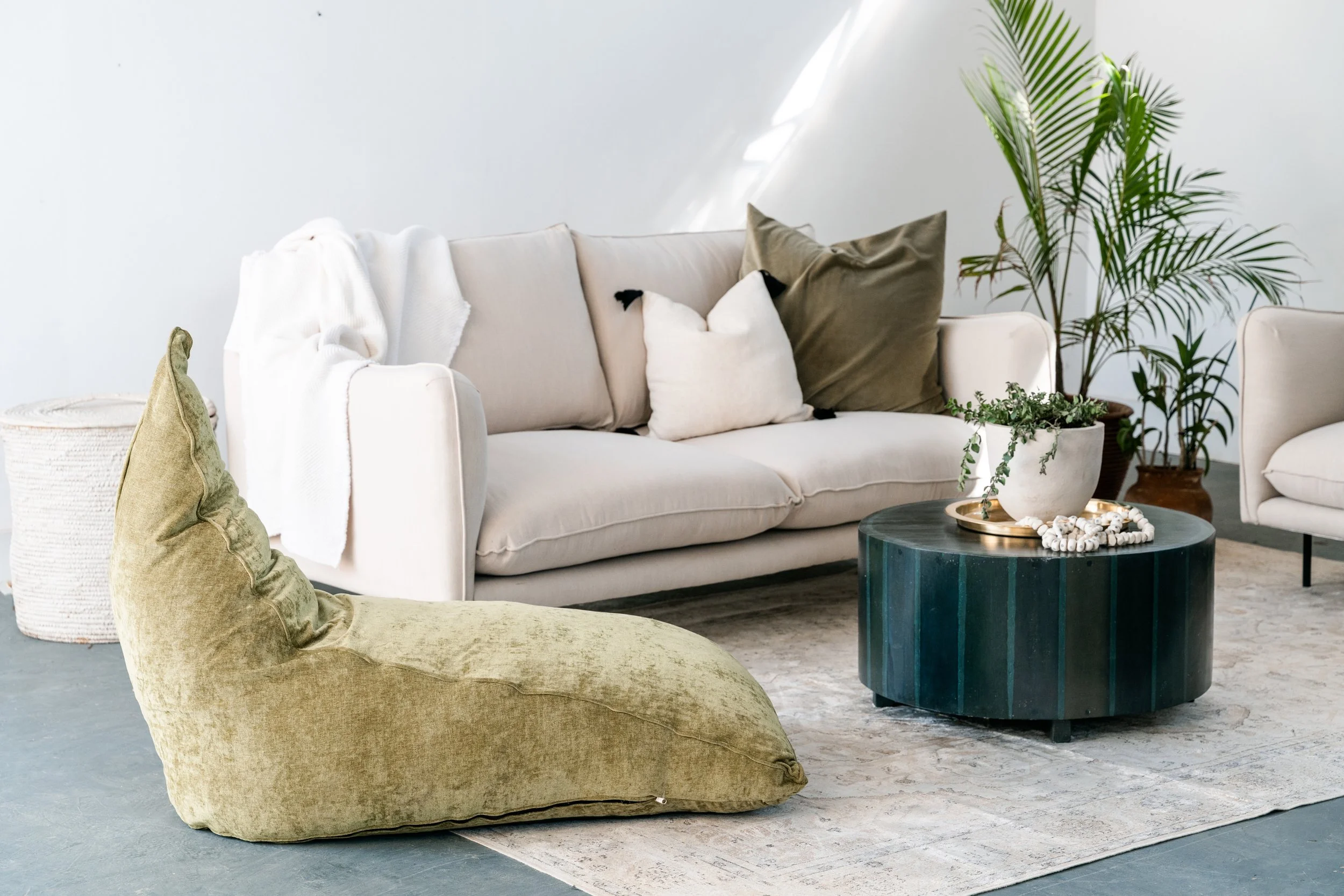 A modern living room with a white sofa accented by multiple pillows, including beige and white ones with black accents. A large leafy green plant stands in a brown pot to the right of the sofa, and a smaller potted plant is on a blue marble coffee table in front of it, which is decorated with a gold tray and white beads. A pale green beanbag is placed on a gray rug on the floor in the foreground.