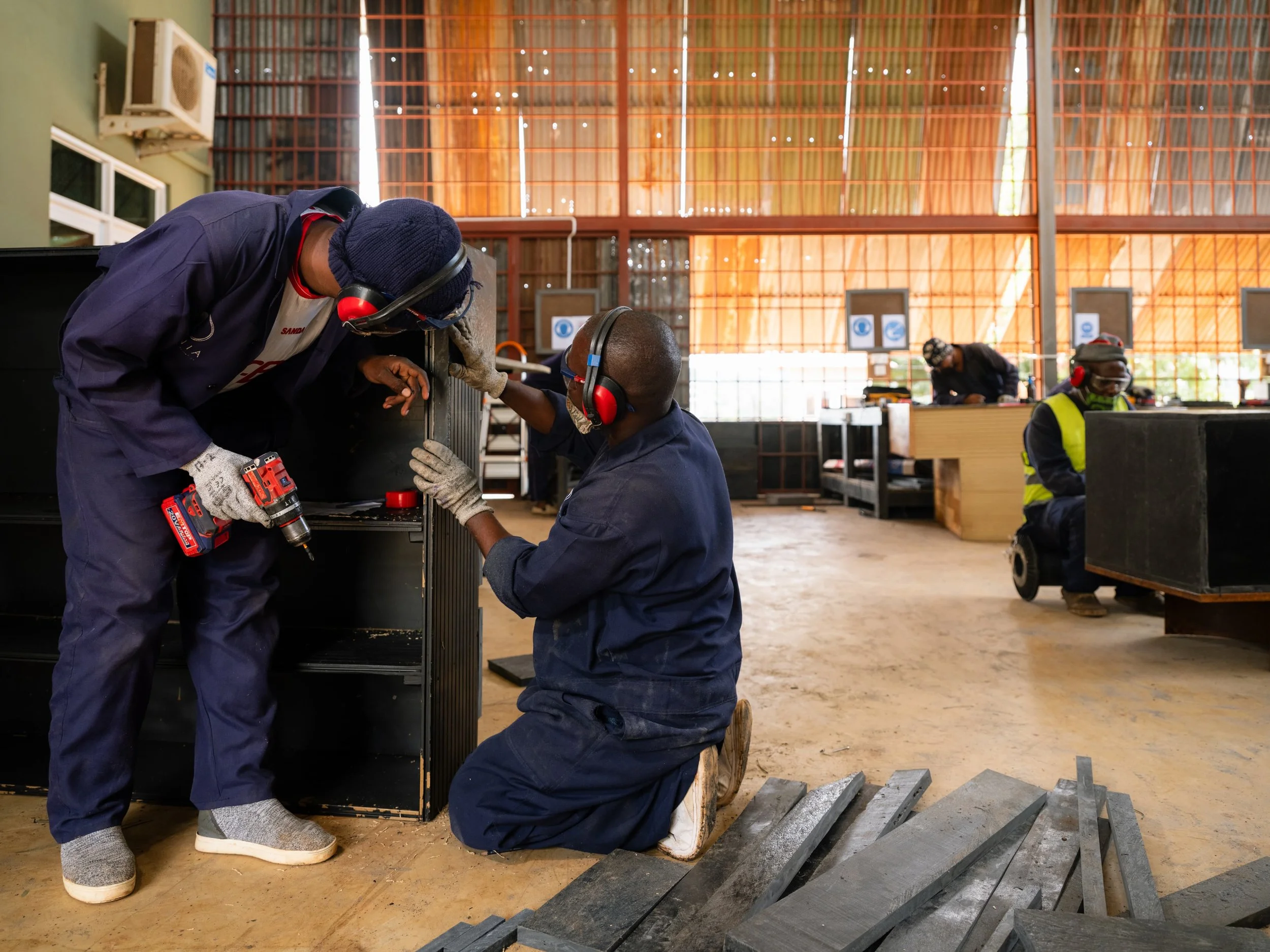 Two workers in a workshop are assembling a piece of furniture or equipment. One is kneeling while the other is standing, both wearing blue uniforms and protective headsets. The standing worker holds a power drill, and the kneeling worker appears to be helping or instructing. Several metal pieces are arranged on the floor nearby, and additional workers can be seen in the background working at desks or stations.