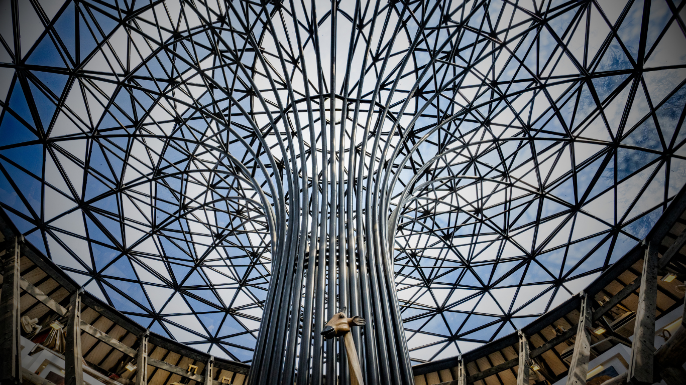 Interior view looking up at a metallic tree sculpture with multiple steel pipes forming the trunk and a geometric dome skylight overhead, revealing partly cloudy sky.