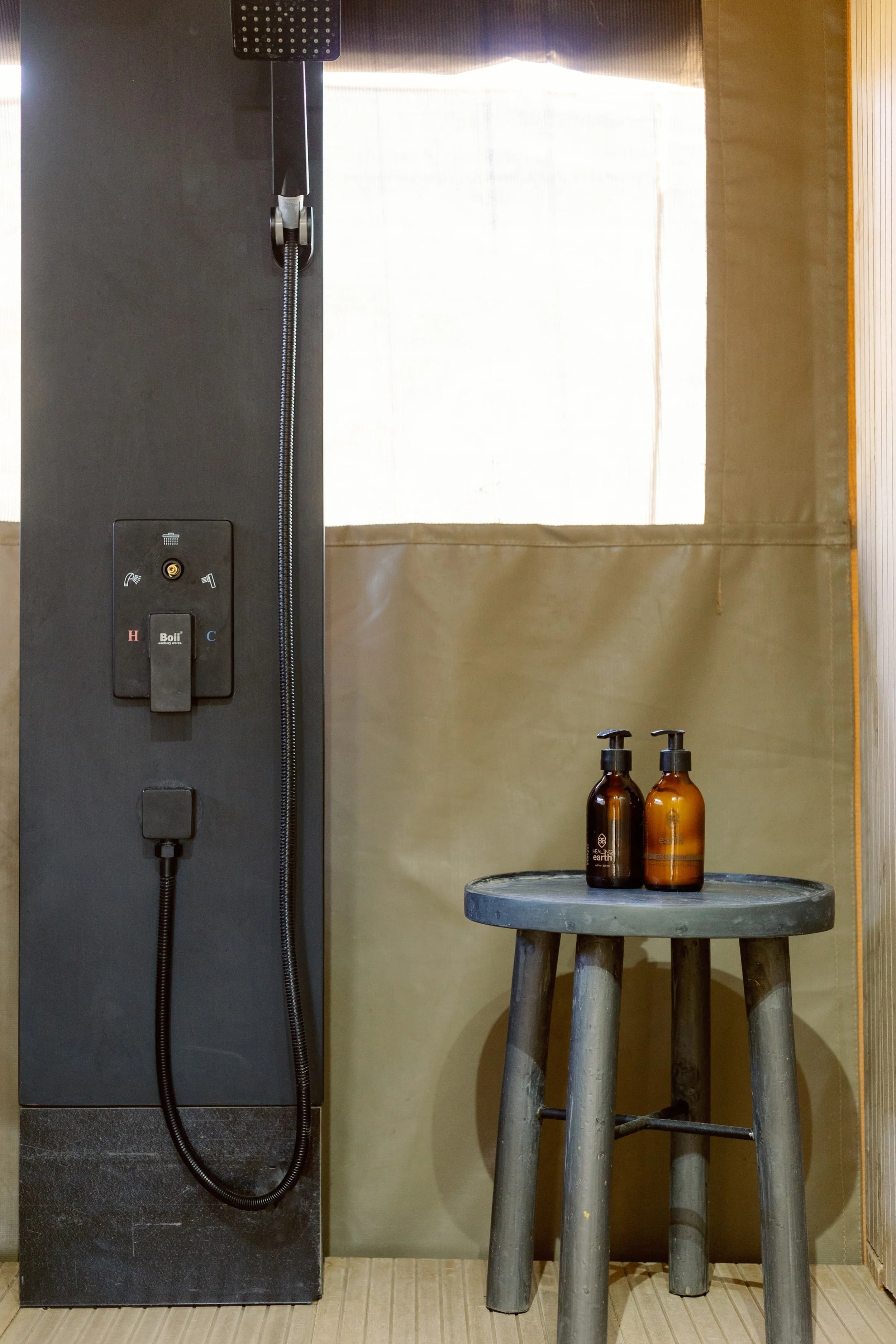 A shower panel with a black finish, featuring controls for hot and cold water and a showerhead at the top, next to a round wooden stool with two amber bottles on top, set against a wall with a beige curtain or fabric.