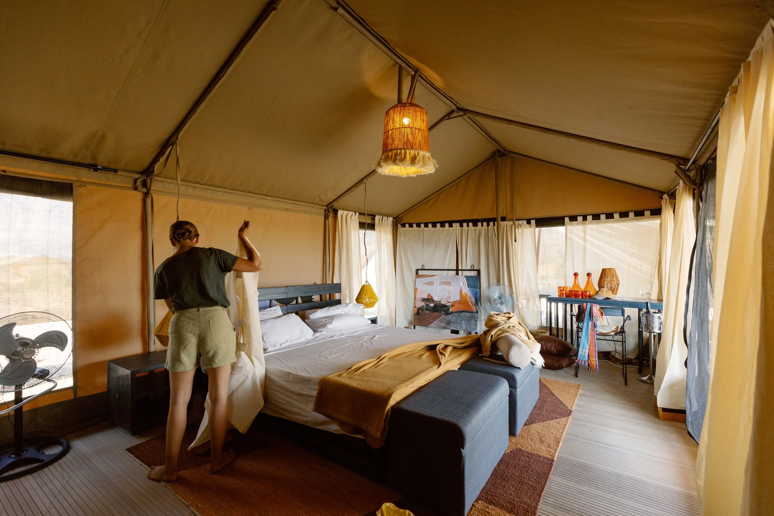 Interior of a tent with a bed, hanging lamp, and a person standing near the bed adjusting the curtains. There are windows with curtains, a fan, and various decorative items on a side table.