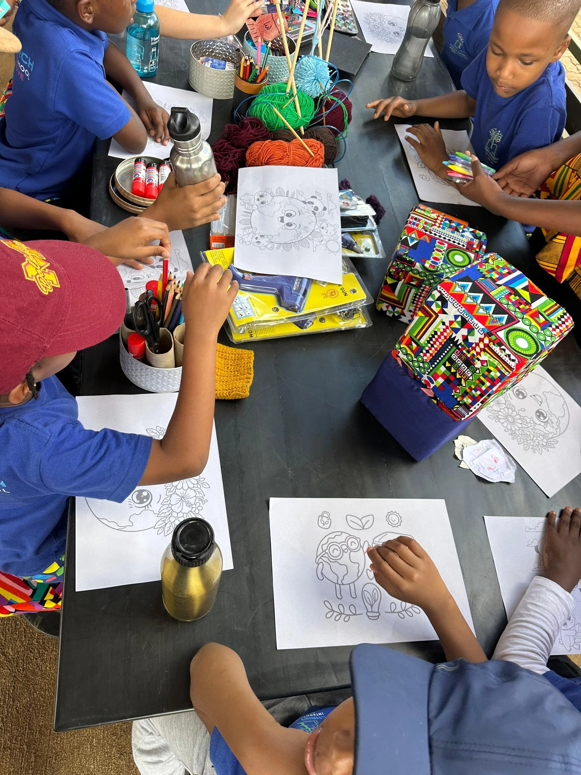 Children sitting at a table coloring and drawing on printable sheets with various crayons and colored pencils. The table has art supplies, yarn, and water bottles. The children are wearing casual clothing including blue shirts and a maroon cap.